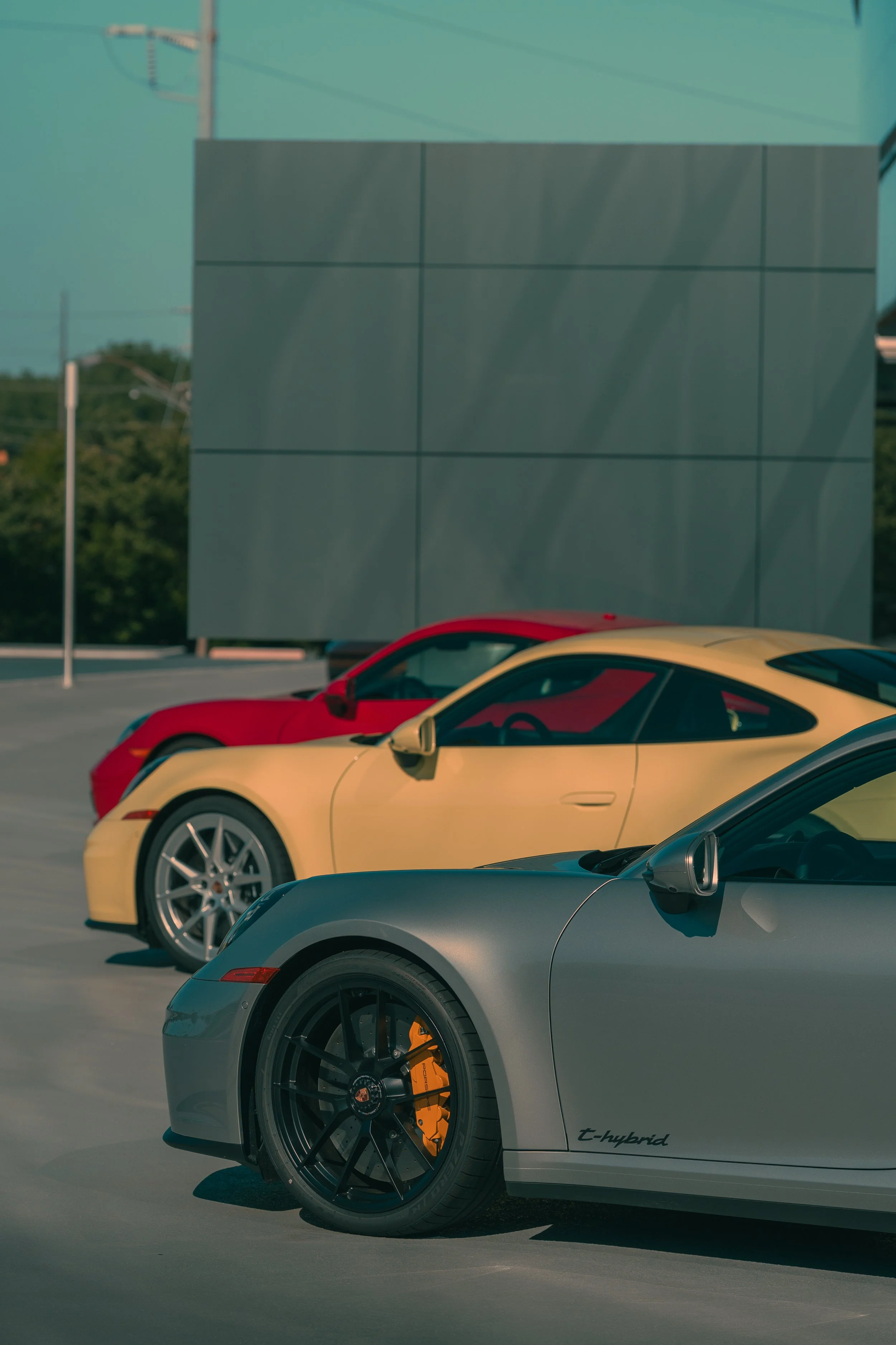 Close-up of a silver Porsche T-hybrid sports car with orange brake calipers in a parking lot, with two other sports cars in red and yellow colors in the background, and a large gray building and trees behind.