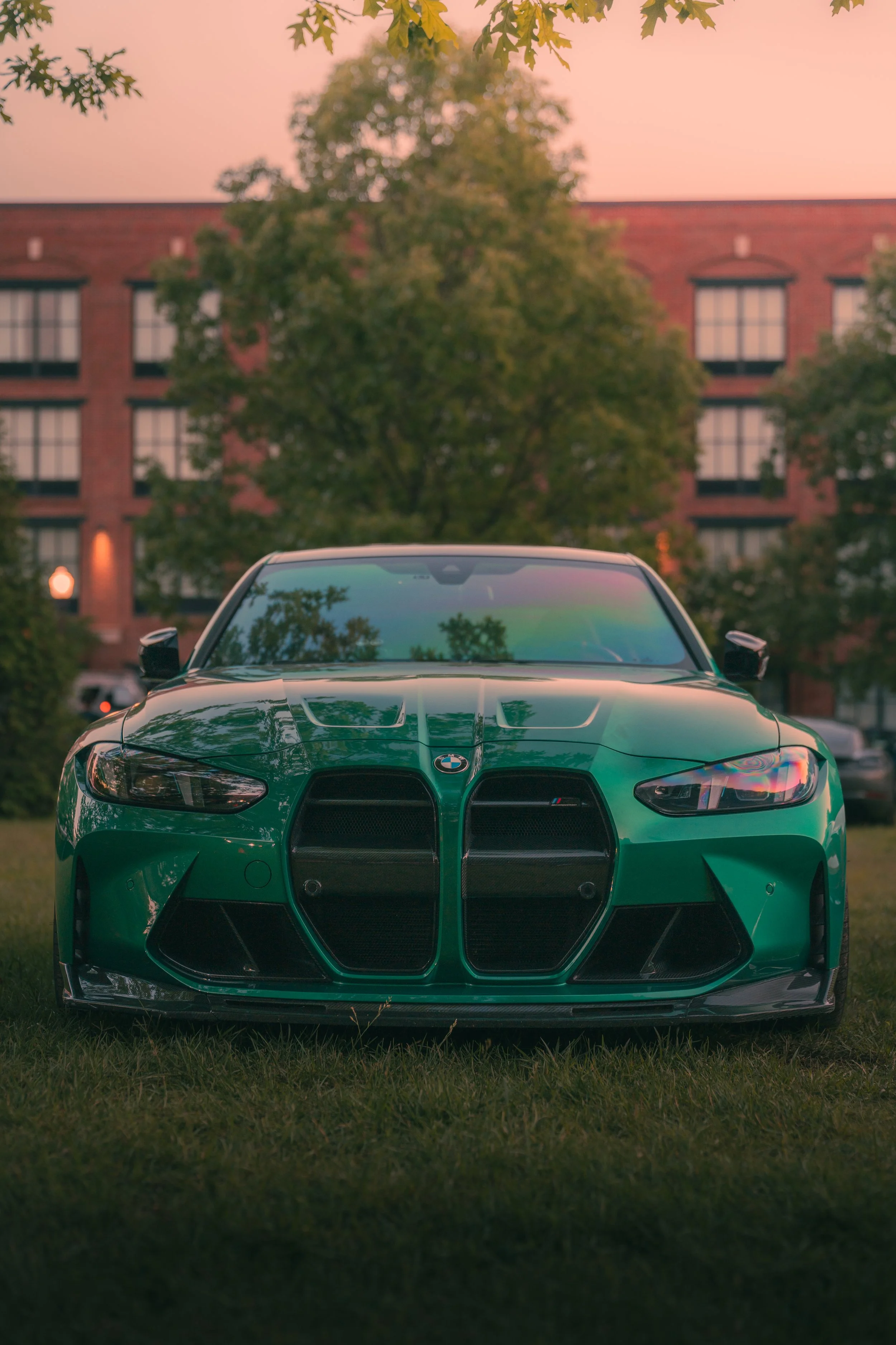 A green sports car parked on a grassy area in front of a red brick building during sunset, with trees on either side and reflections of trees on the car's windshield.