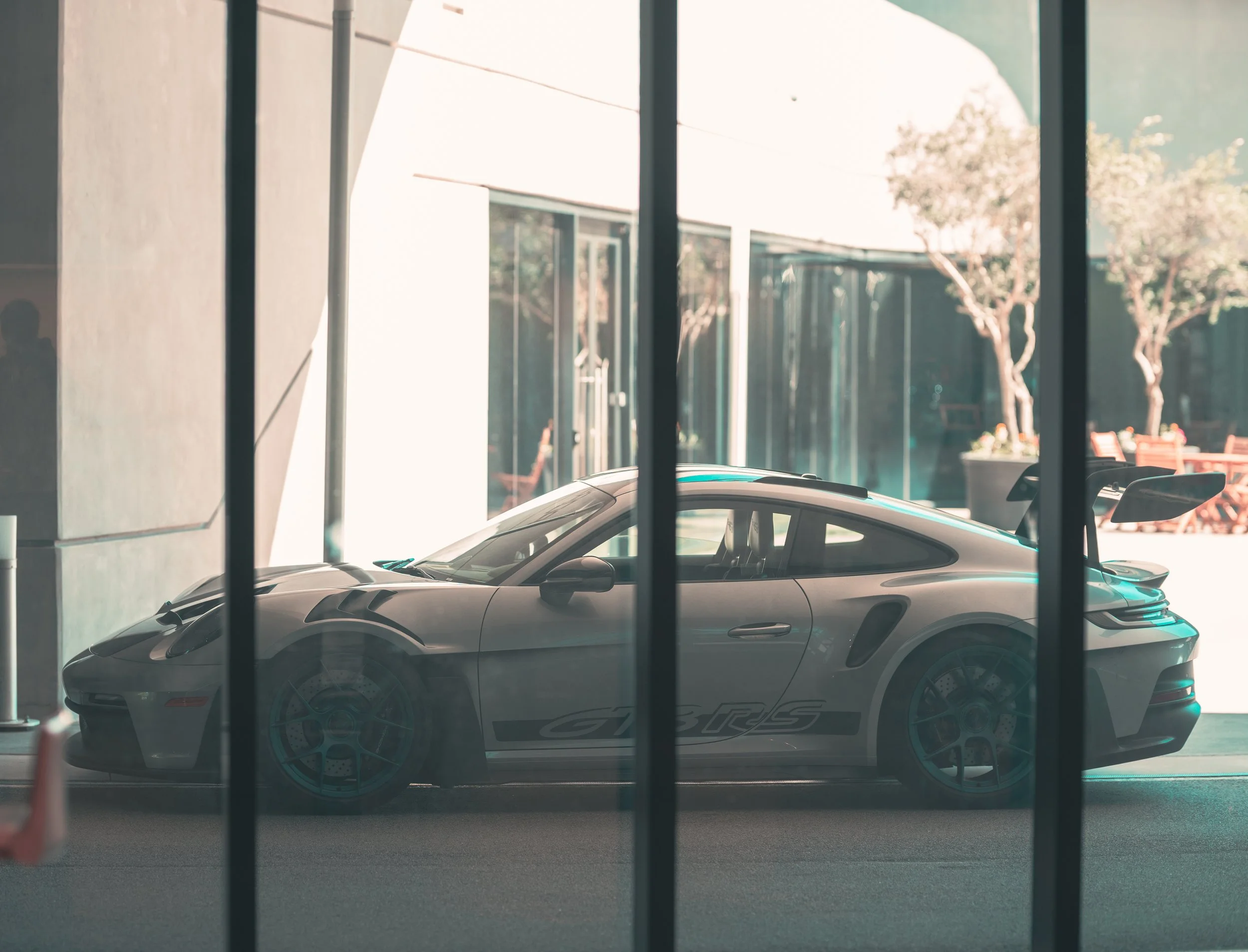 View of a silver sports car with a spoiler inside a building, photographed through a window with black frames; outdoor trees and orange chairs are visible in the background.
