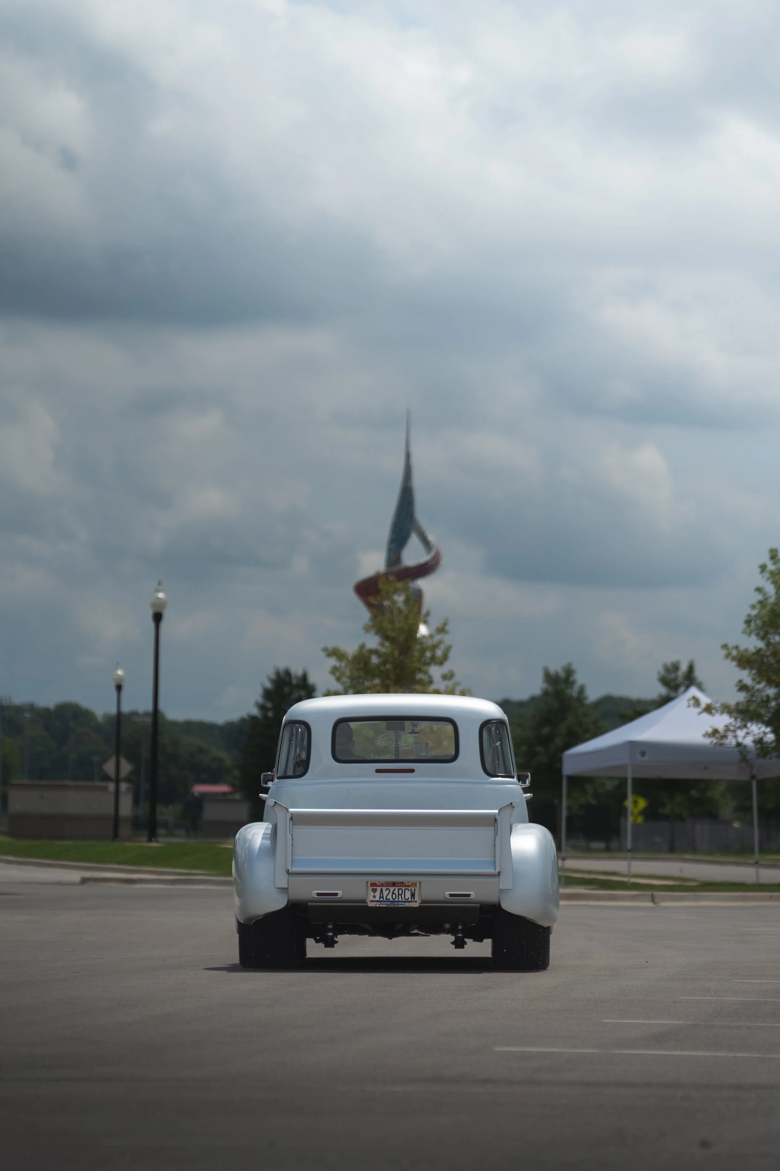 A vintage white pickup truck facing away in an open parking lot under a cloudy sky with dark clouds, with trees, lampposts, and a tent in the background.