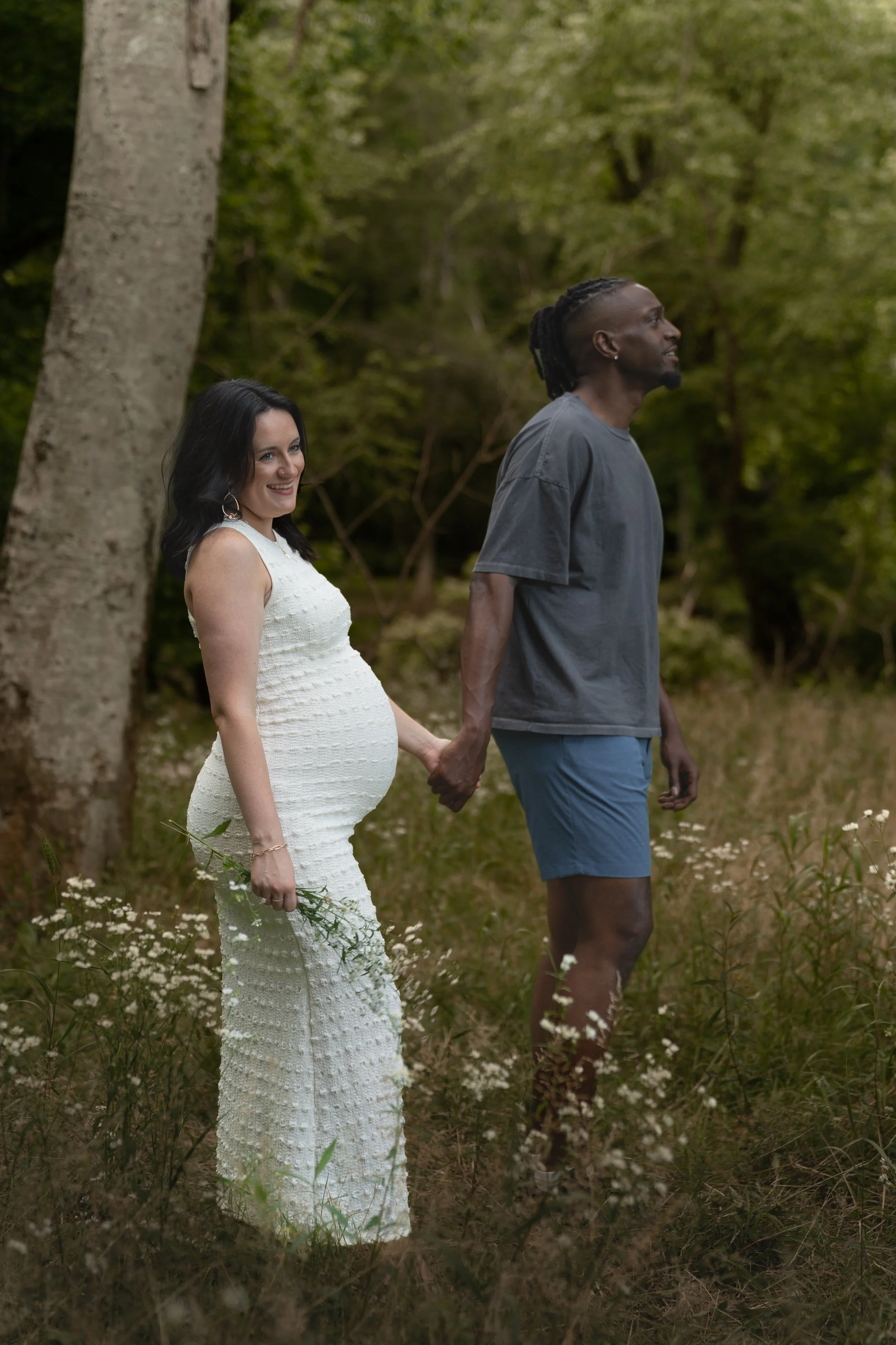 A pregnant woman in a white dress holding flowers and smiling, standing hand-in-hand with a man in a grey T-shirt and blue shorts, in a forest with green trees and wildflowers.