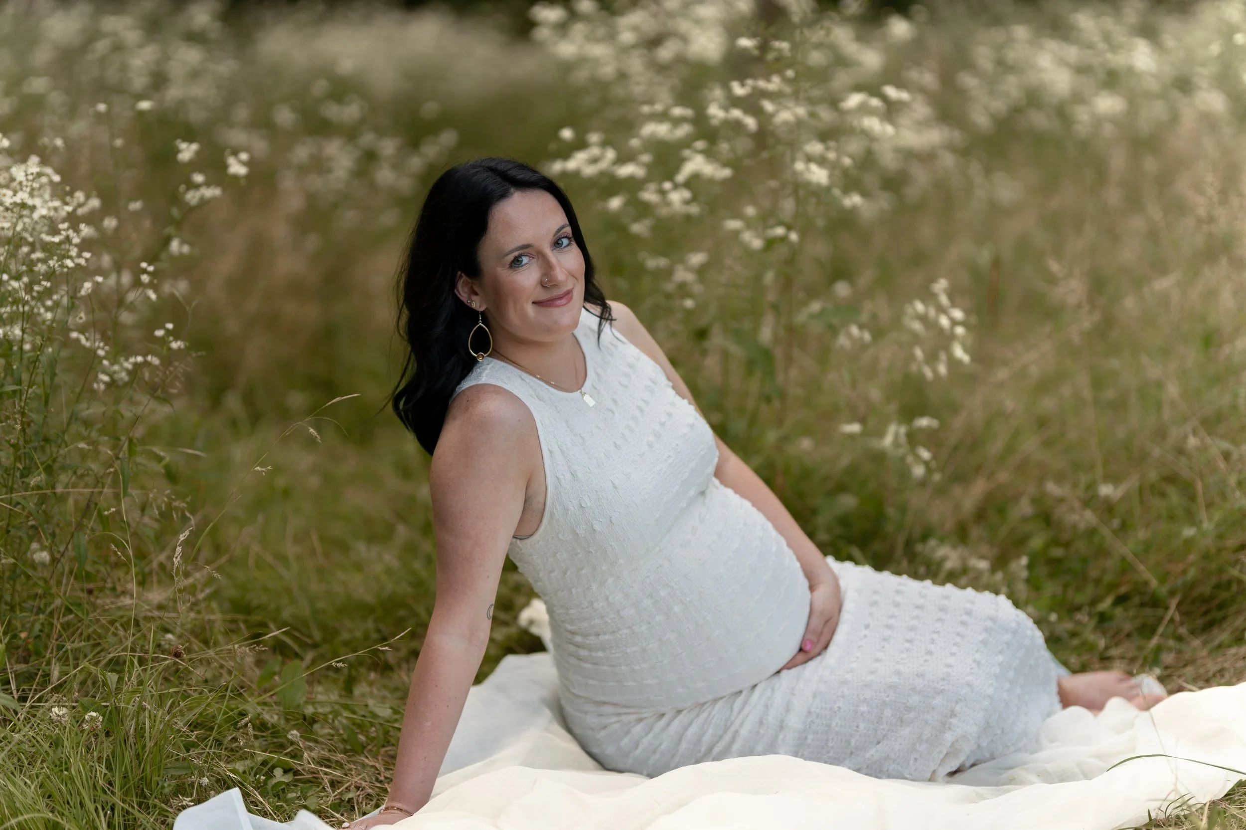 A pregnant woman with black hair and blue eyes, wearing a white dress, sitting outdoors on a white blanket in a grassy area with small white flowers.