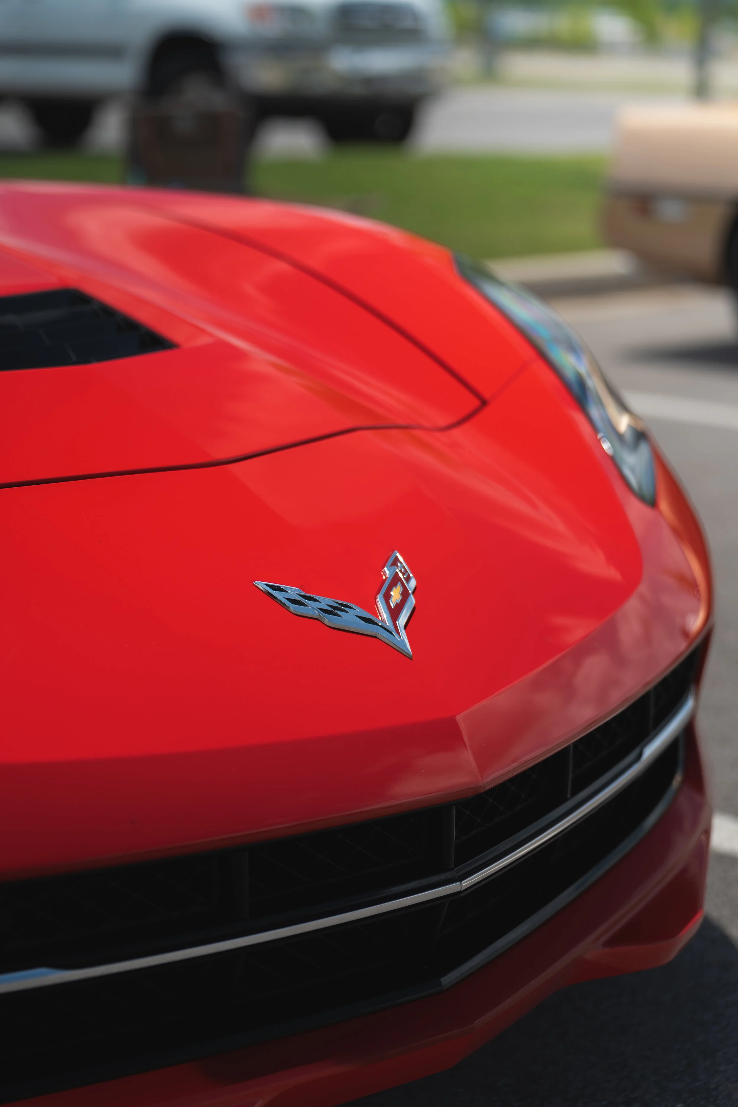 Close-up of the front of a red Chevrolet Corvette sports car with the Chevrolet logo and a Corvette emblem on the hood, parked outdoors near other vehicles.
