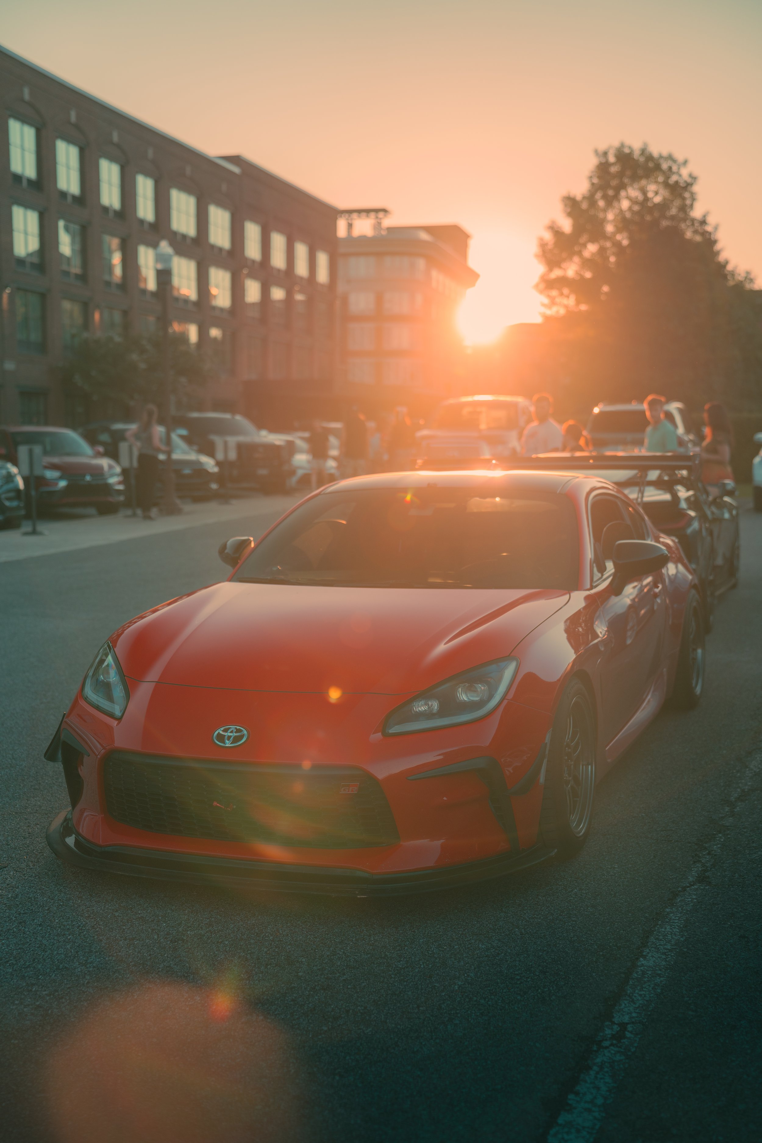 A red Toyota sports car parked in a lot during sunset with a building and several people in the background.