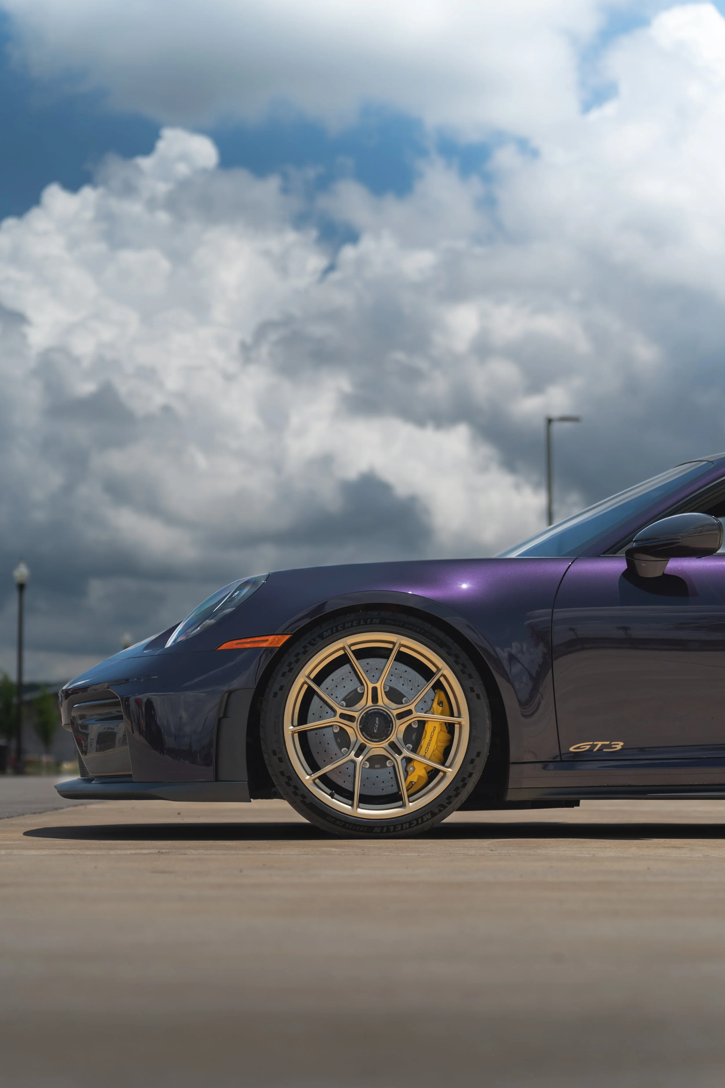 Partial view of a dark blue Porsche 911 GTR3 sports car with golden wheels, yellow brake calipers, and a sleek design, parked outdoors under cloudy skies.