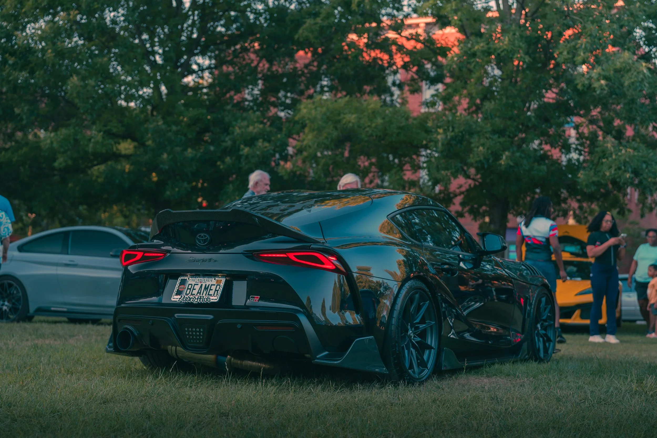 Black Toyota Supra sports car parked on grass with trees and people in the background during sunset.