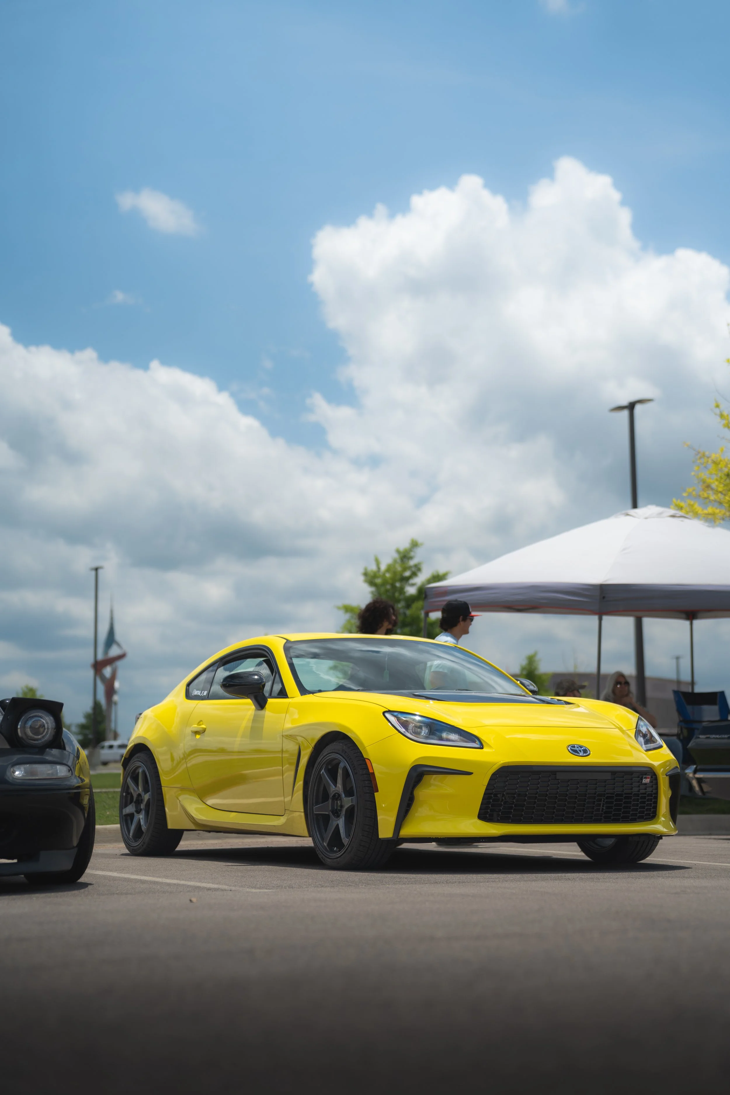A yellow sports car parked in a parking lot at an outdoor event, with a tent and people in the background under a partly cloudy sky.