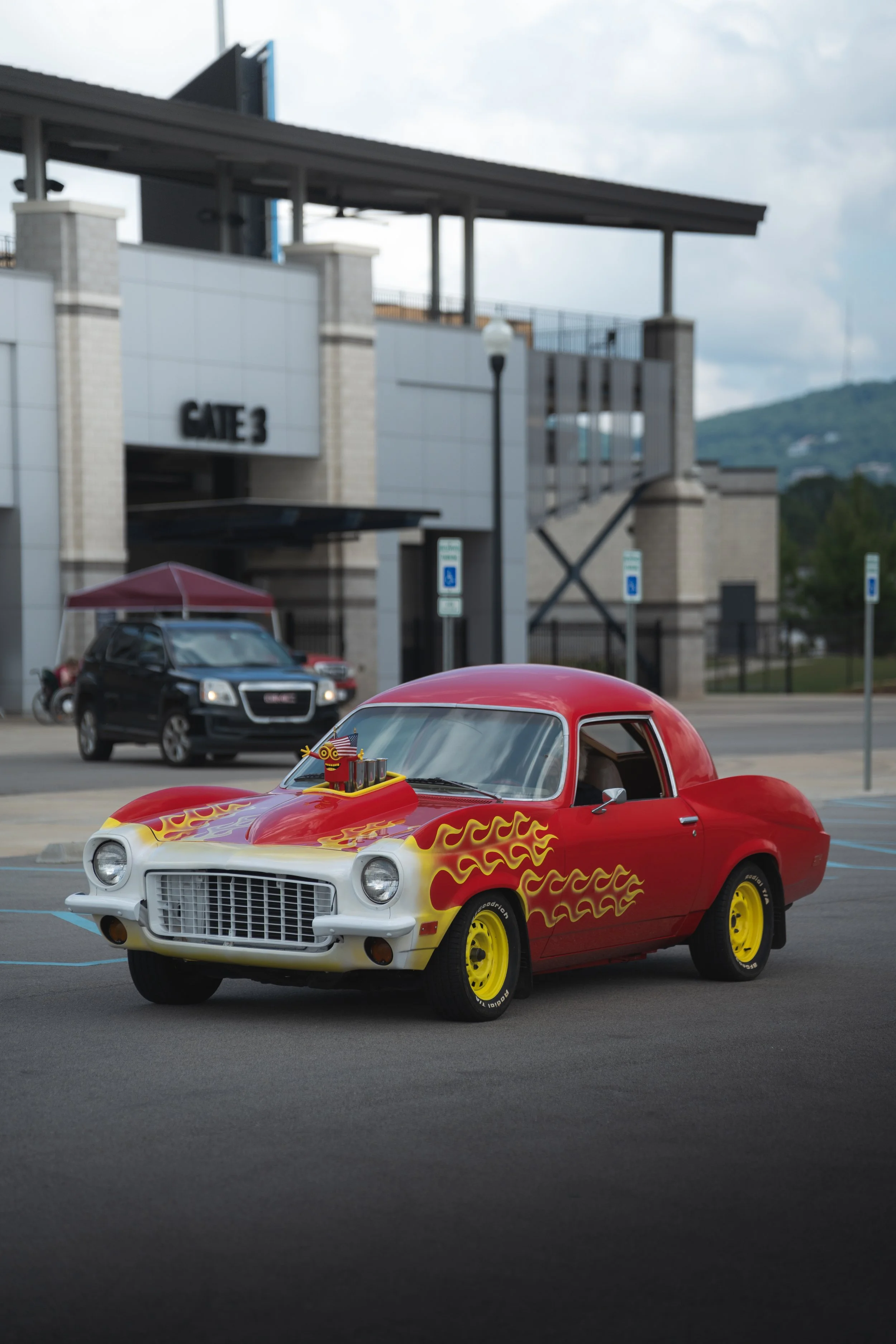 A red and white vintage car with yellow wheels and flames painted on its sides, parked in a parking lot with a building in the background.
