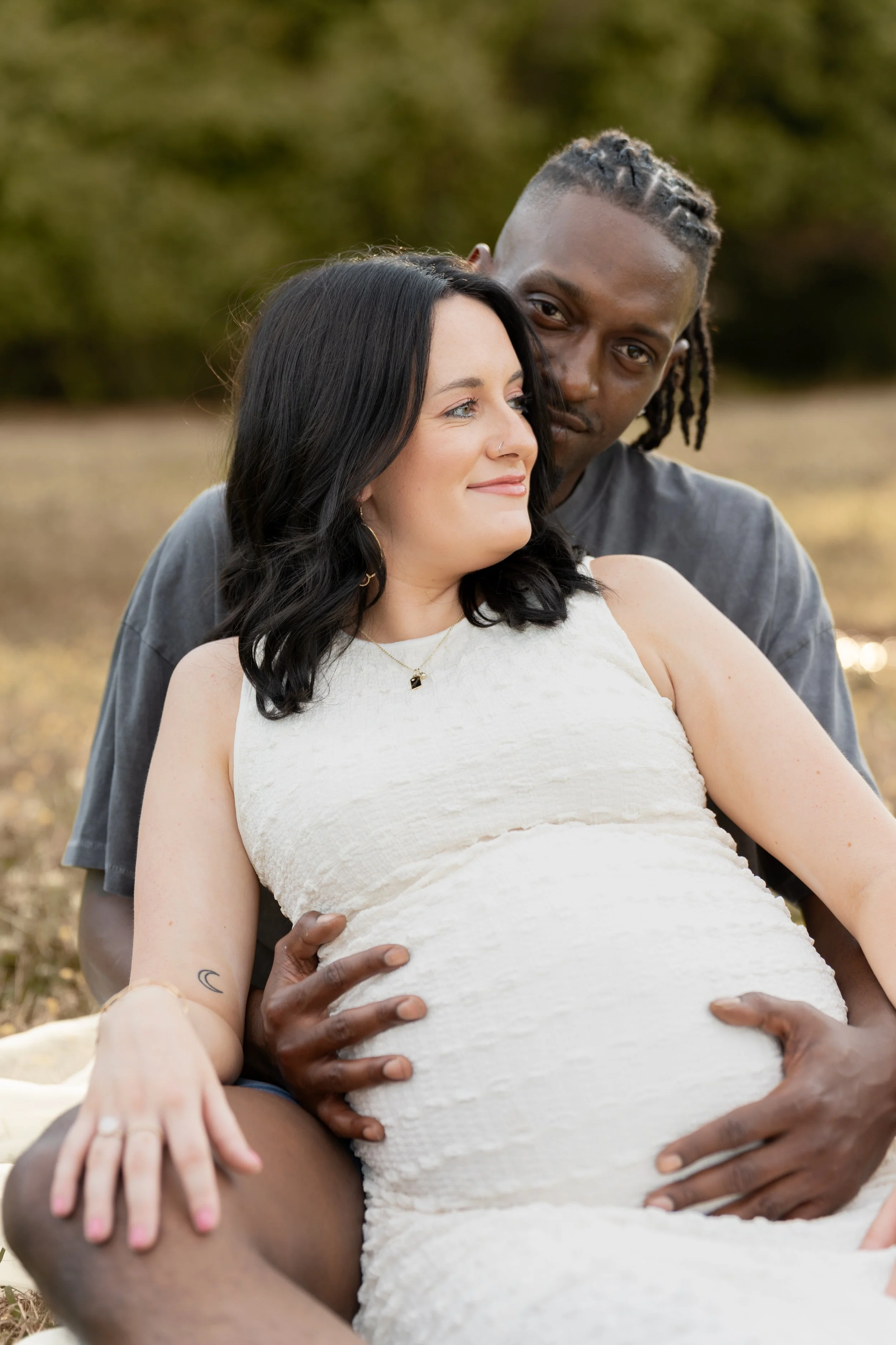 A diverse couple sitting outdoors, with the woman in a white dress and the man behind her. They are smiling and holding her pregnant belly, with the man's hands resting on her stomach. The background shows blurred outdoors scenery.