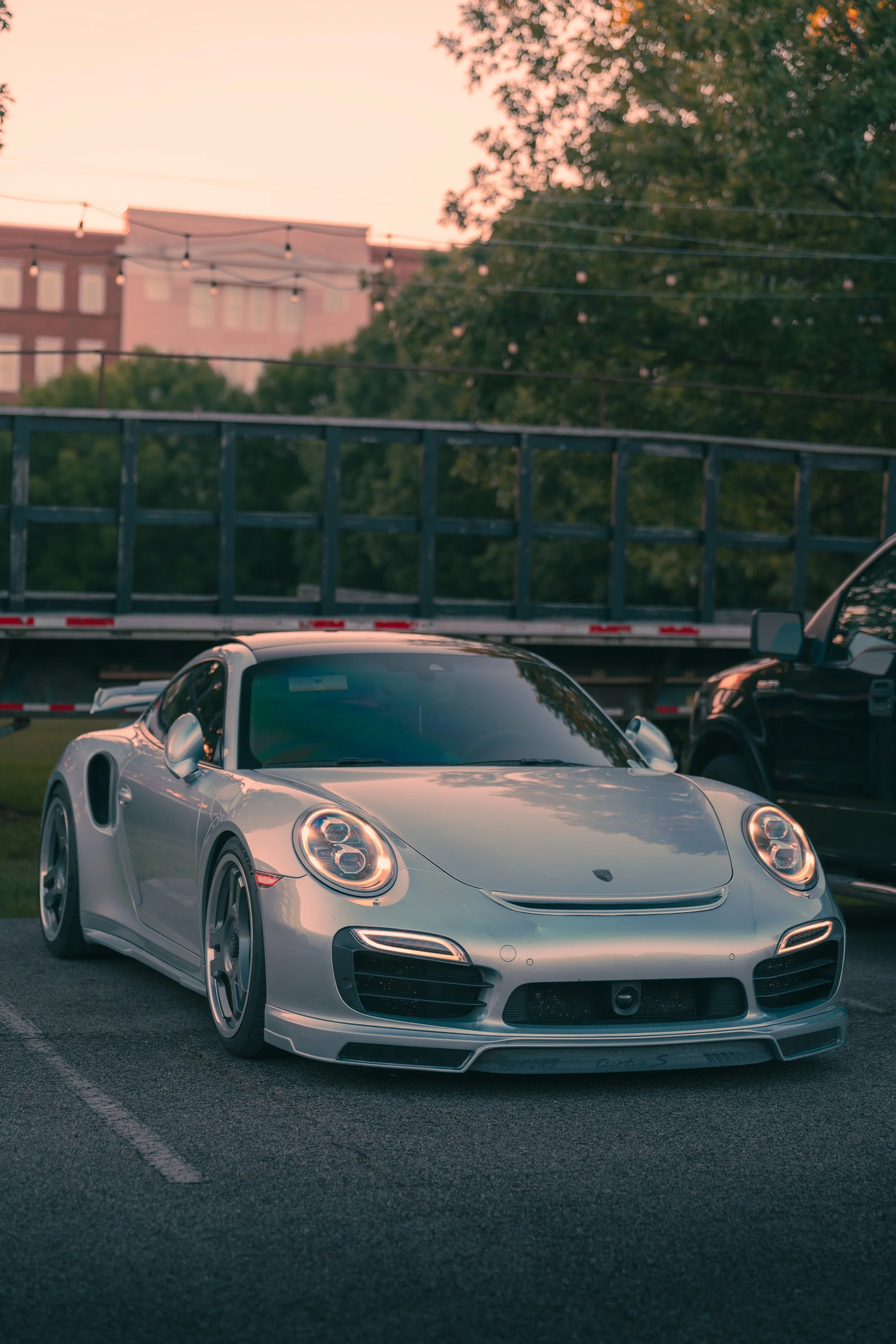 A silver Porsche 911 Turbo S sports car parked in a parking lot during dusk, with a bridge and trees in the background.