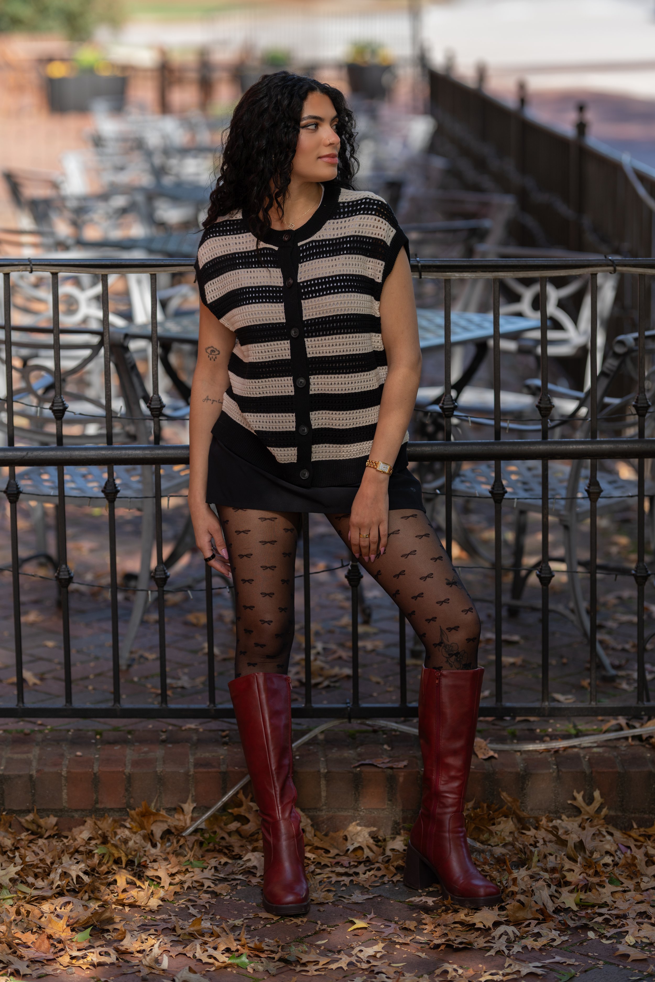 Young woman with curly dark hair wearing a striped black and beige top, sheer black stockings with bow patterns, red boots, and a wristwatch, standing outdoors on a fall day with fallen leaves, metal tables and chairs, and a railing in the background.
