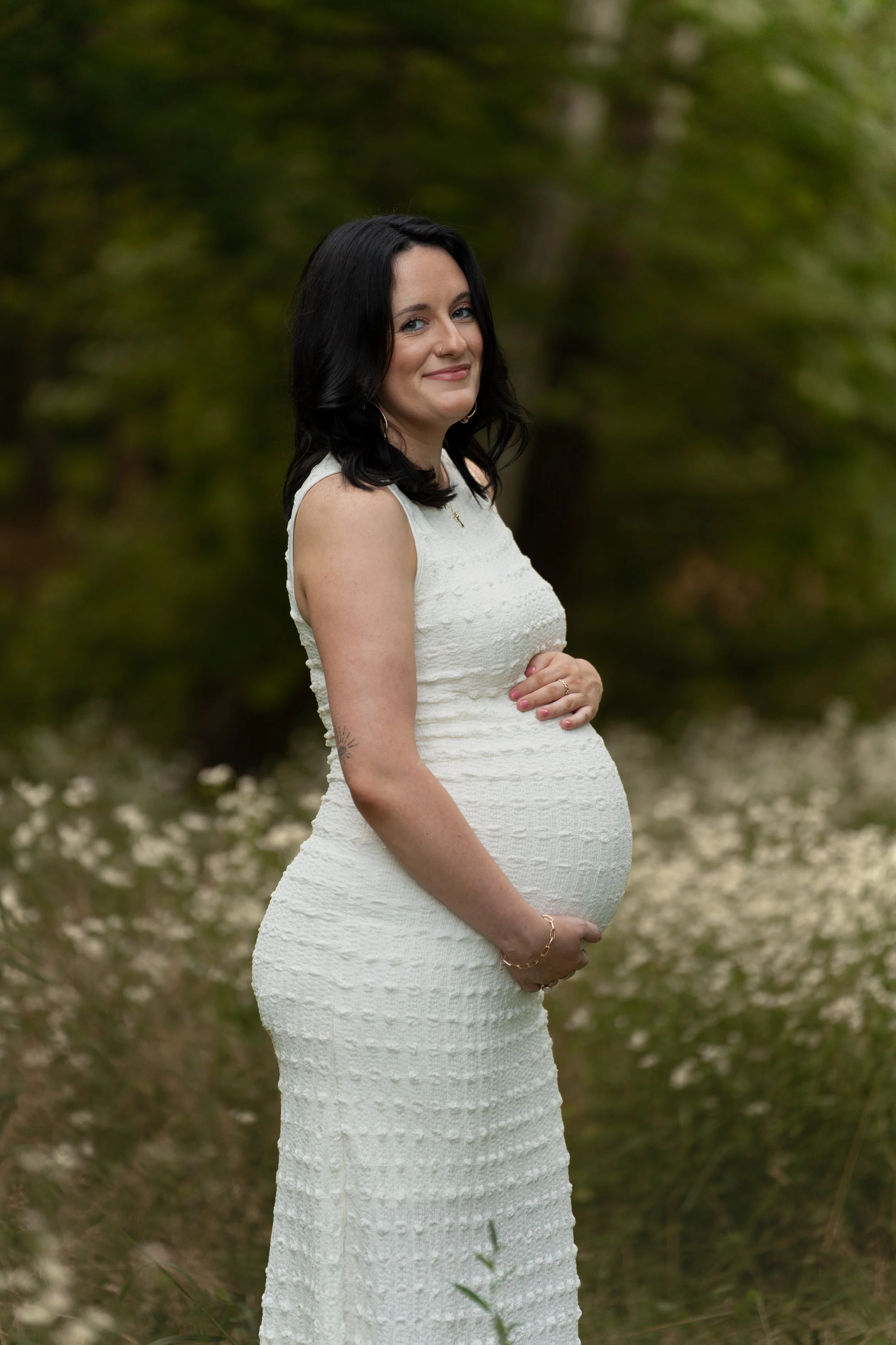 A pregnant woman with black hair in a white textured dress standing outside in a natural area with green trees and white flowers on the ground, smiling gently and holding her belly.