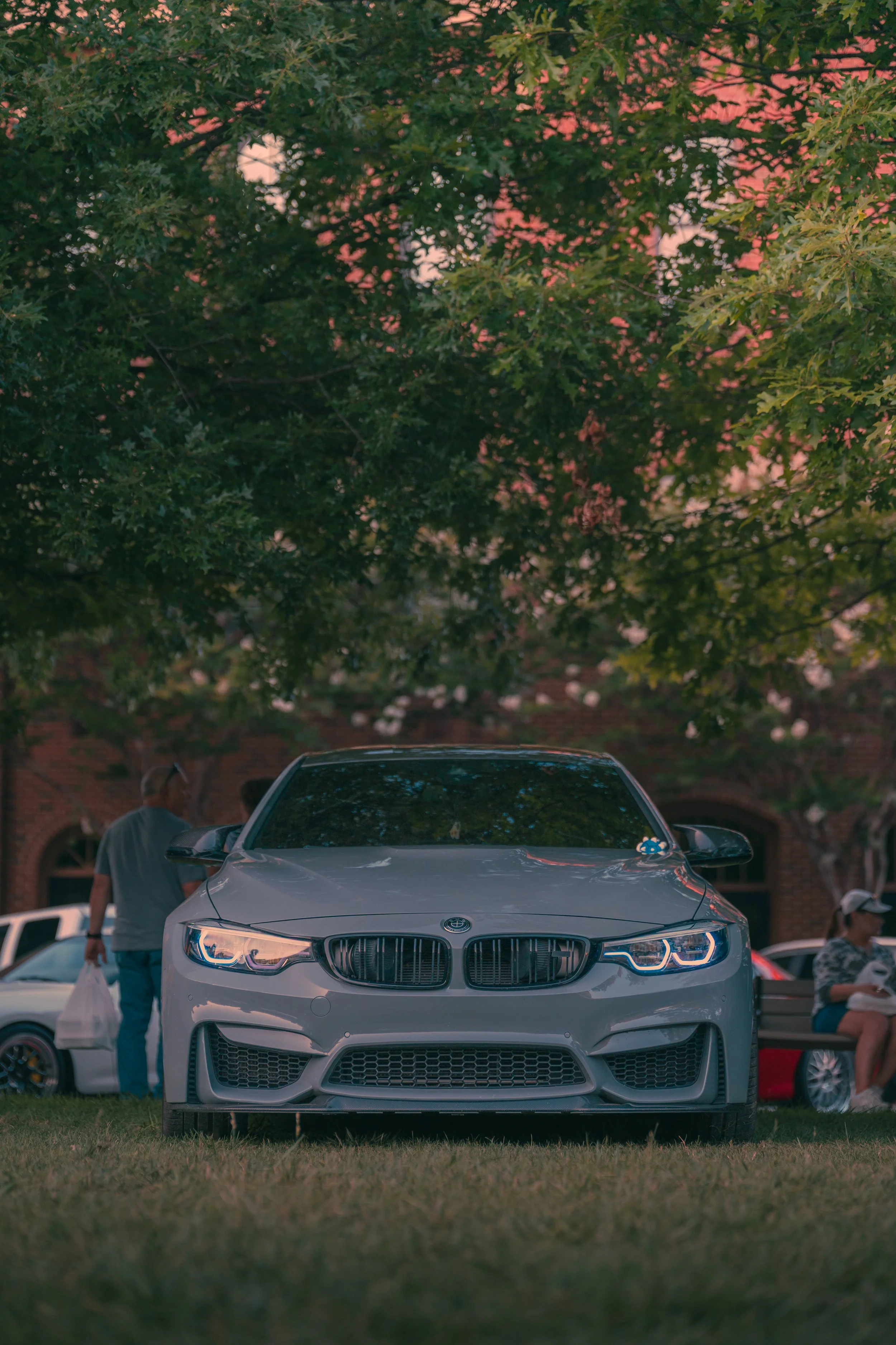 A white BMW car parked on grass in front of a brick building with green trees in the background. People are visible near the car, one holding shopping bags.