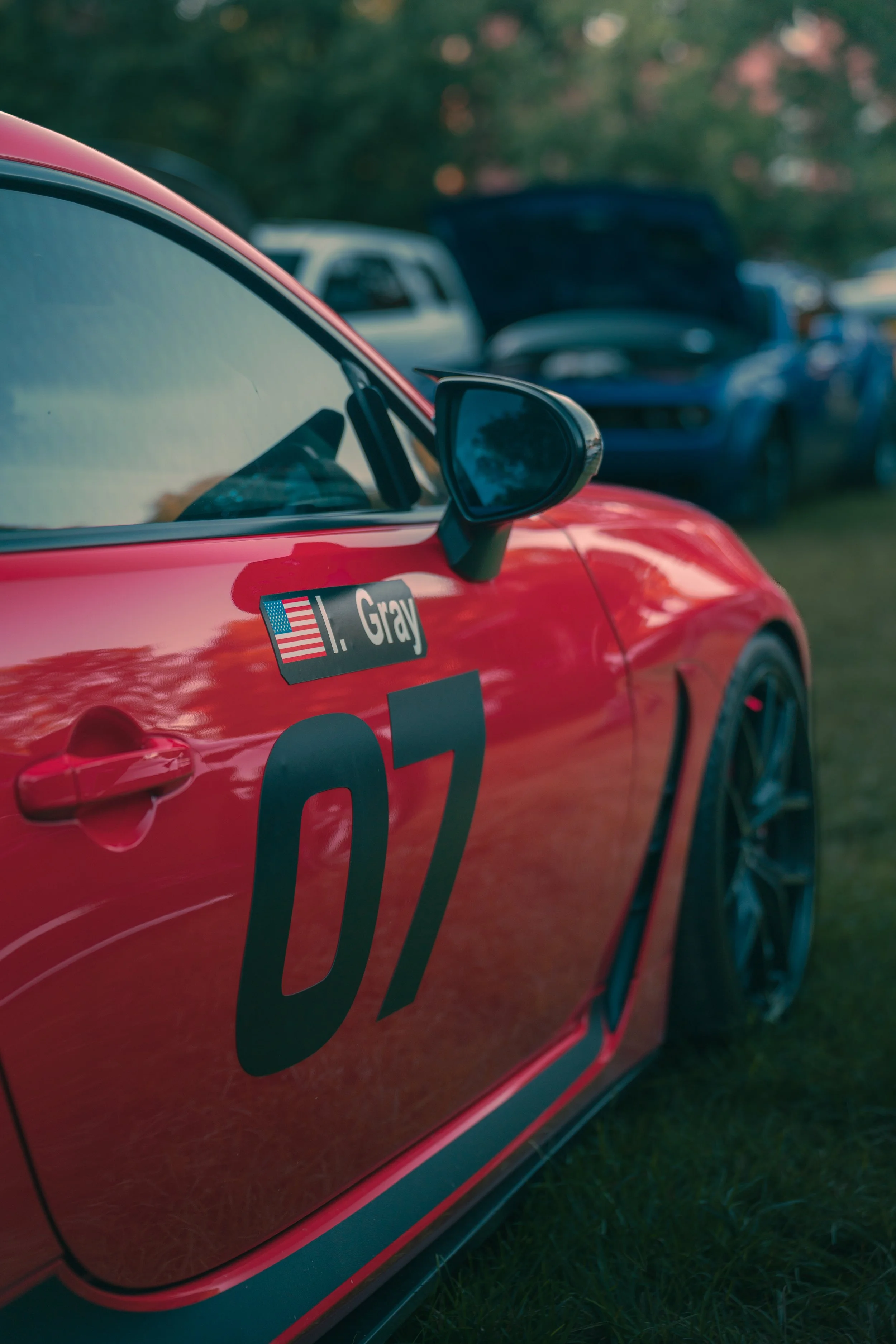 Close-up of a red race car with number 97 and a sticker of an American flag, parked on grass, with other vehicles in the background.