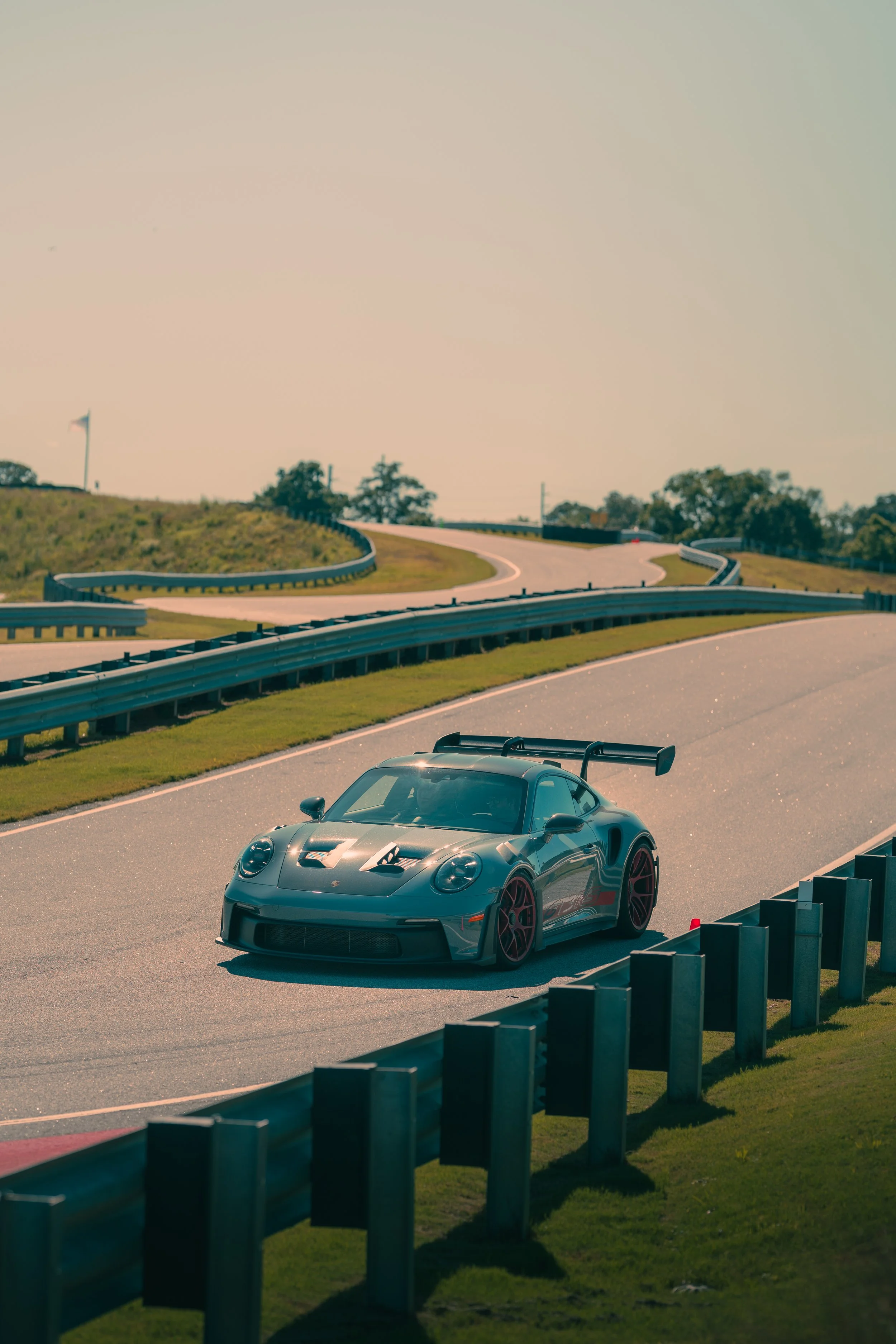 A black sports car with a rear wing is parked on a winding racetrack with safety barriers. The track curves upward into a hill with grass and trees, and a flag is visible in the distance against a clear sky.