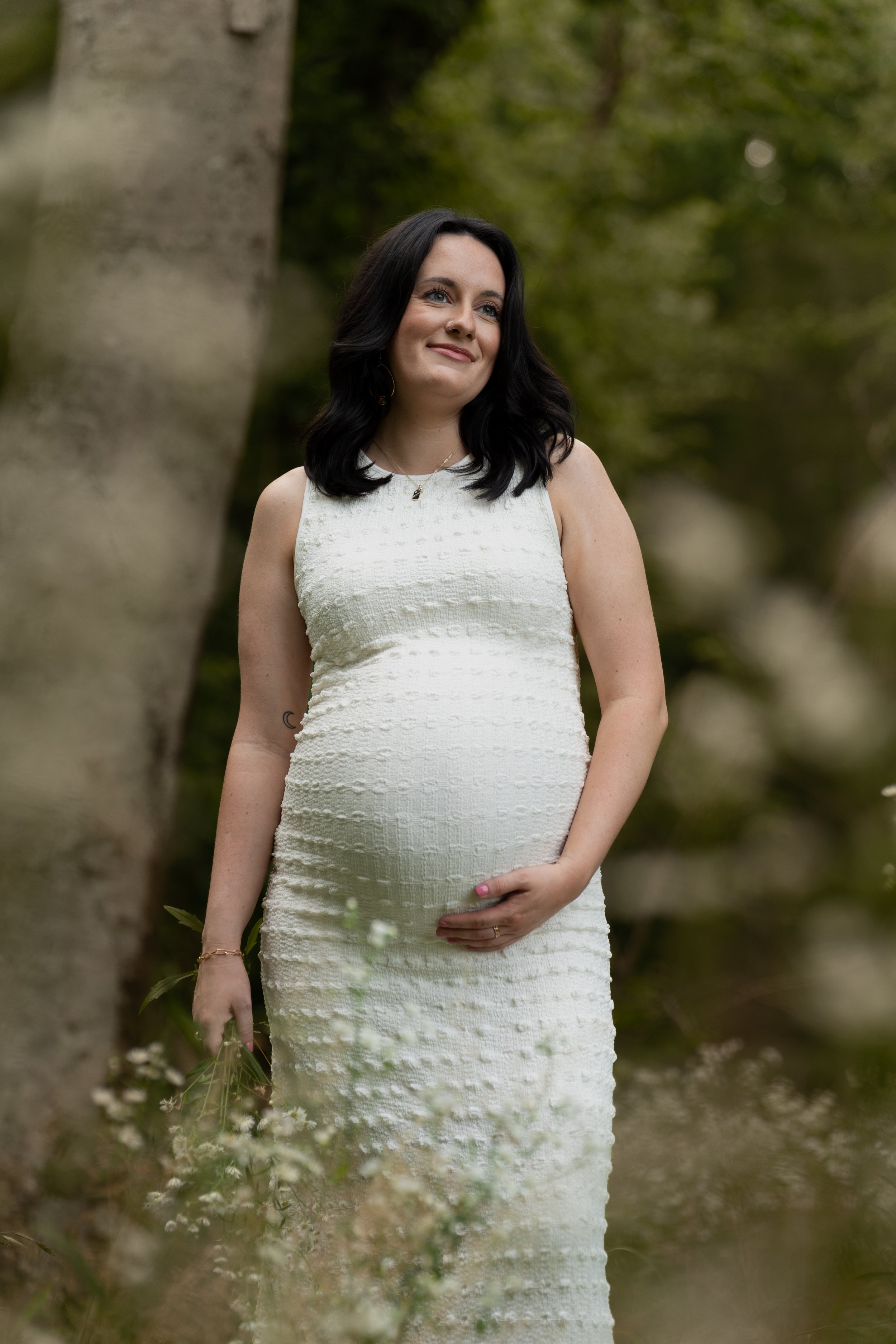 A pregnant woman in a white dress standing outdoors among trees and plants, holding her belly with one hand and looking to the side with a gentle smile.