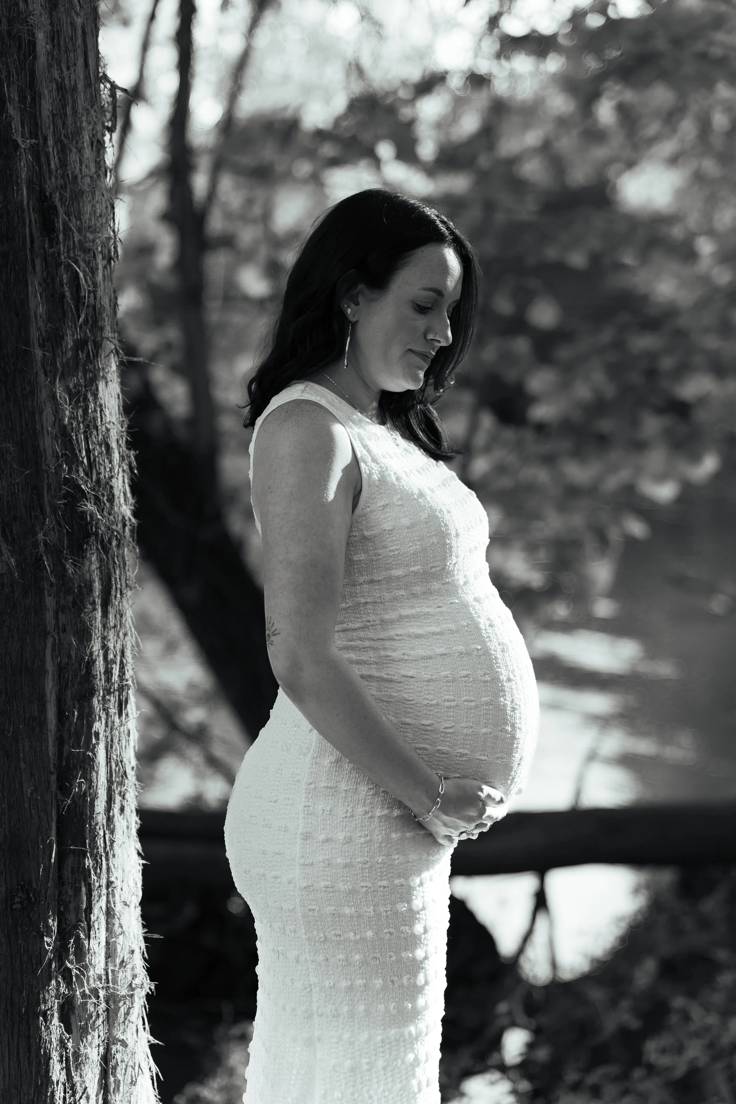 A pregnant woman in a white dress standing outdoors next to a tree, gently holding her belly and smiling softly, with a blurred natural background.