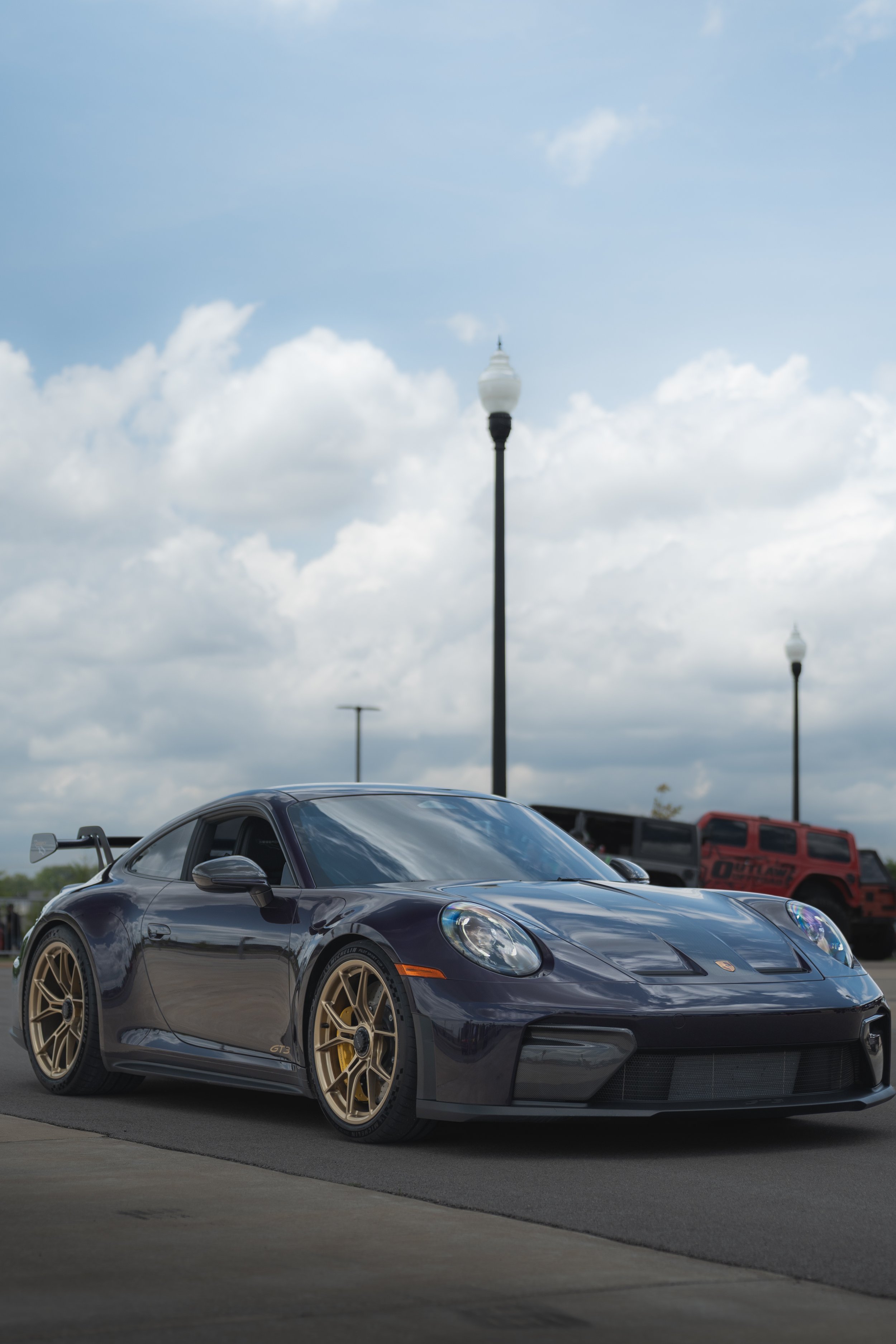 A black sports car with gold wheels and a rear spoiler parked on a concrete surface, with street lamps and vehicles visible in the background under a cloudy sky.