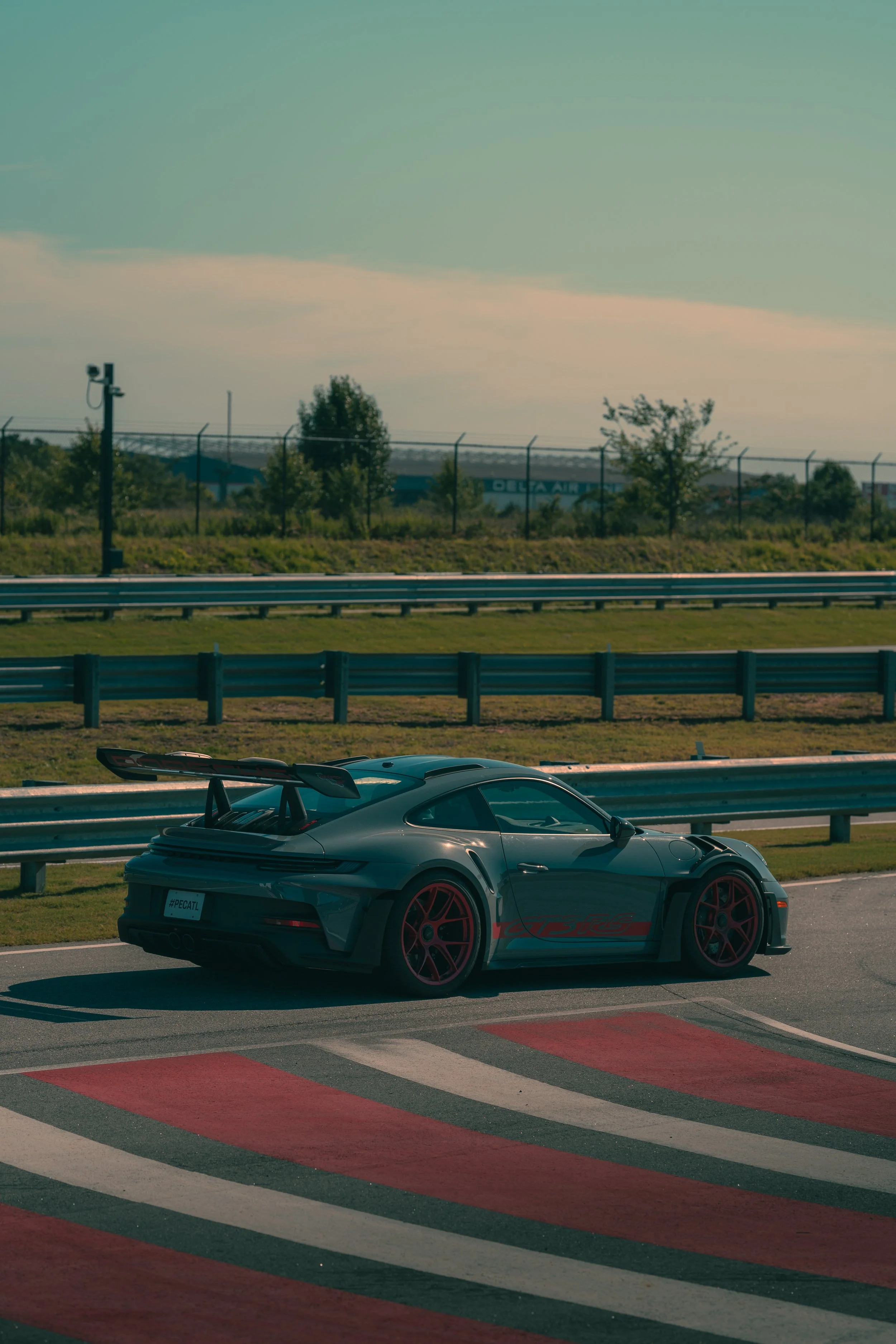 A gray sports car with red accents and a large rear wing, parked on a race track with red, white, and black painted curbs in the foreground, and a fence, trees, and a Delta Air Lines building in the background.