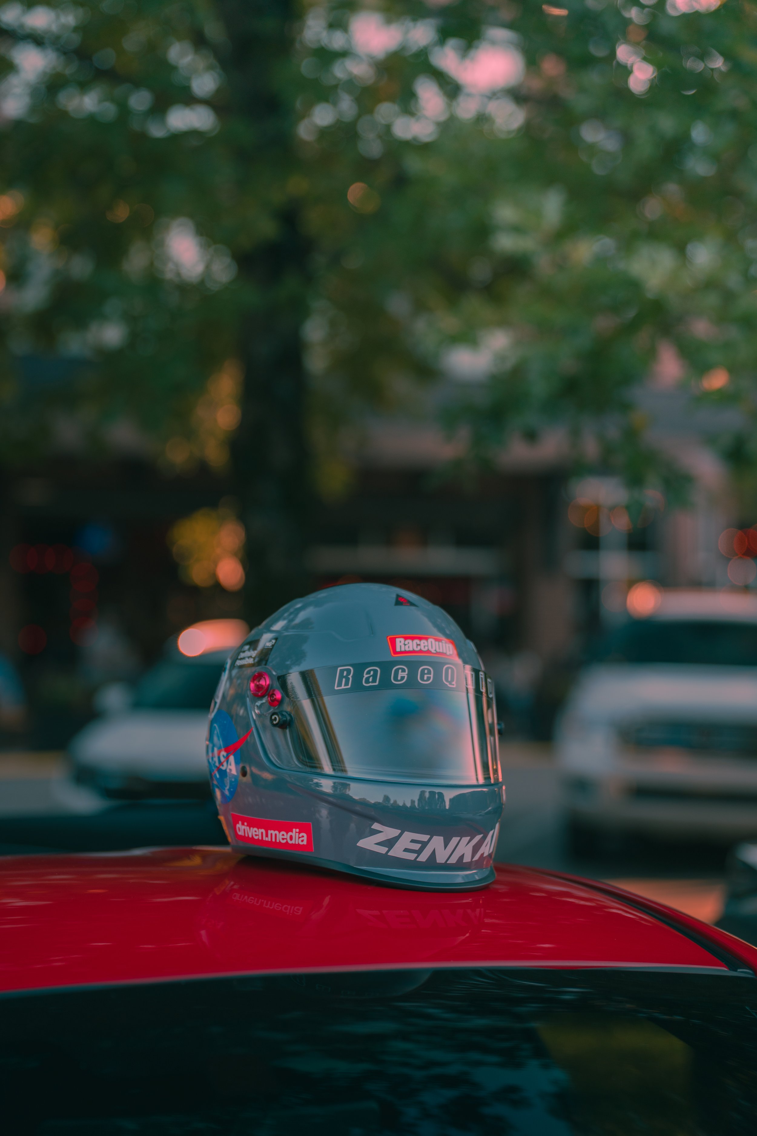 A racing helmet with sponsorship stickers is placed on the roof of a red car. The background is blurred and shows trees and cars, indicating a parking lot or urban scene.
