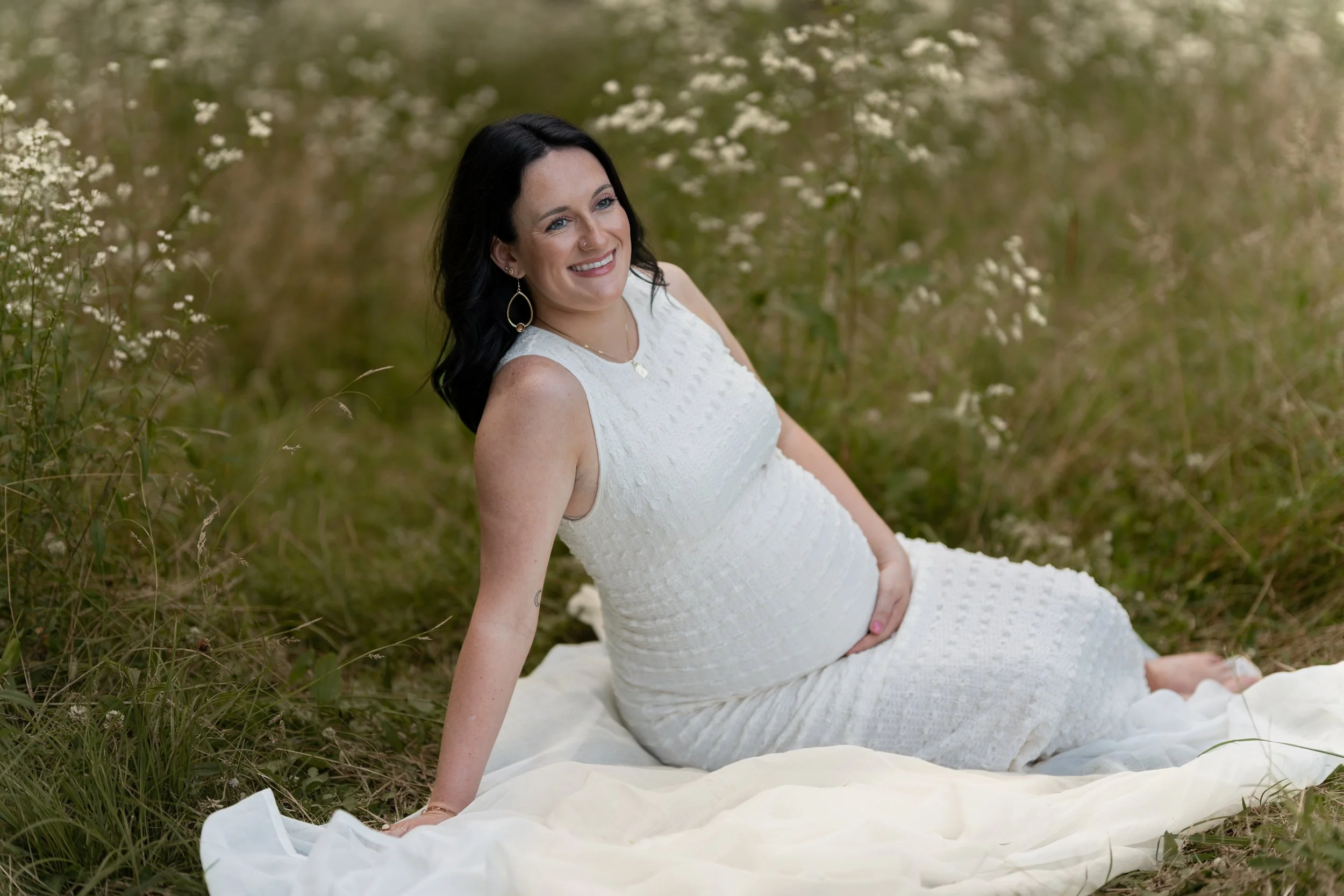 A woman with black hair and a nose piercing, wearing a white lace dress, sitting on a white fabric outdoors in a grassy area with small white flowers, smiling and looking to her left.