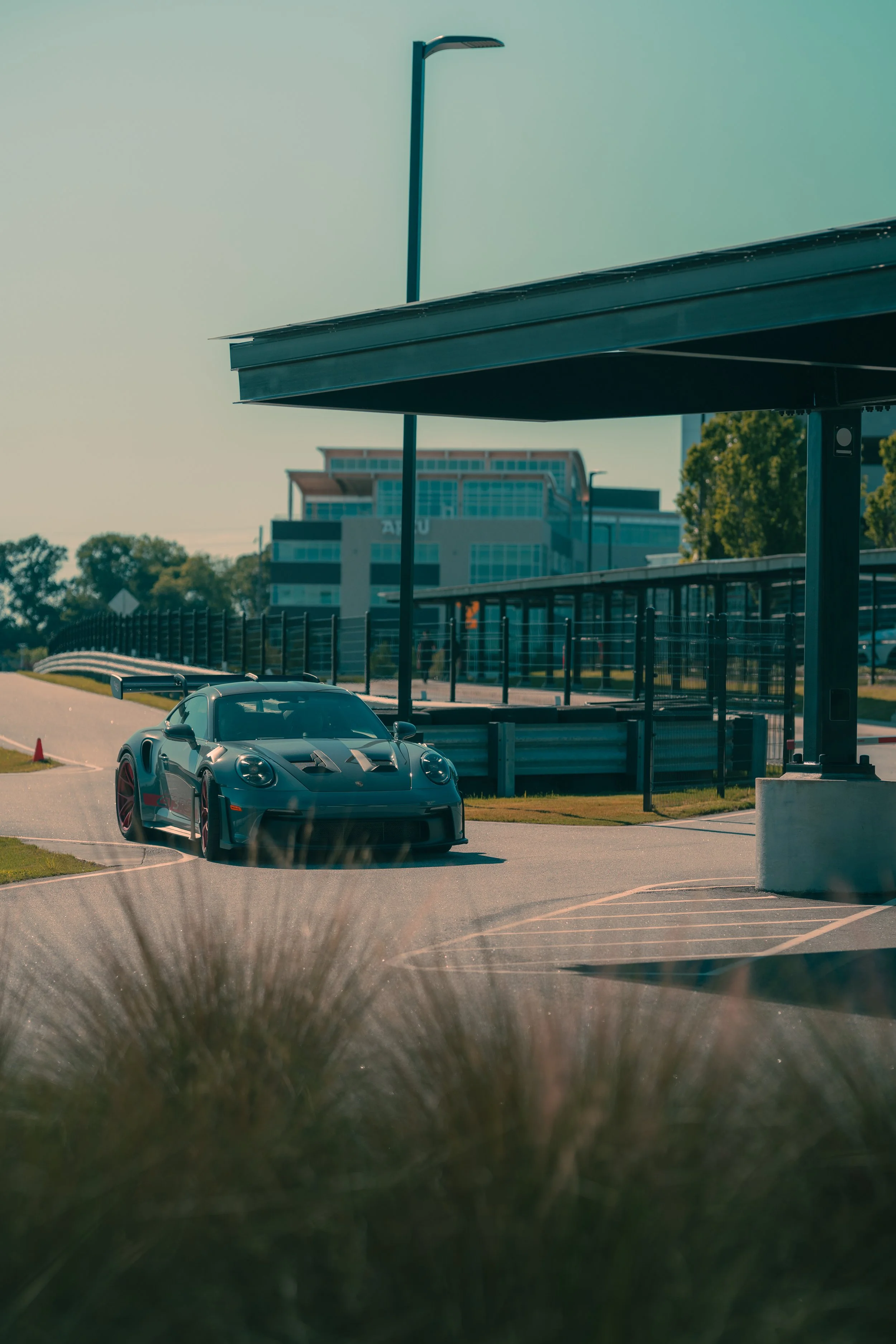 A black sports car with red accents parked near a curb on a sunny day, with a modern building, trees, and a curved bus stop shelter in the background.