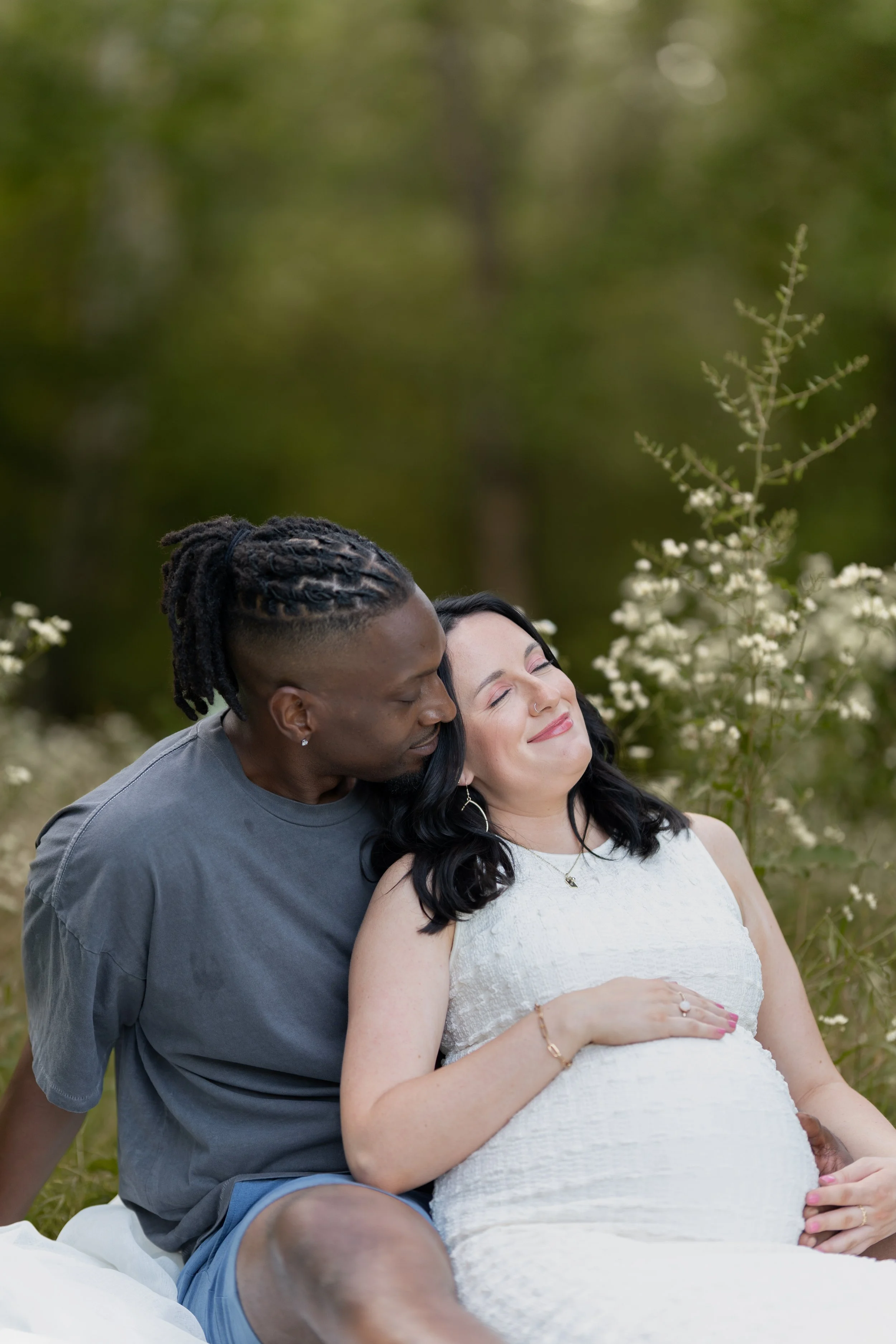 A couple sitting outdoors, the man with dreadlocks and the woman with dark hair and a nose ring, with the woman appearing pregnant, surrounded by white flowers and greenery.