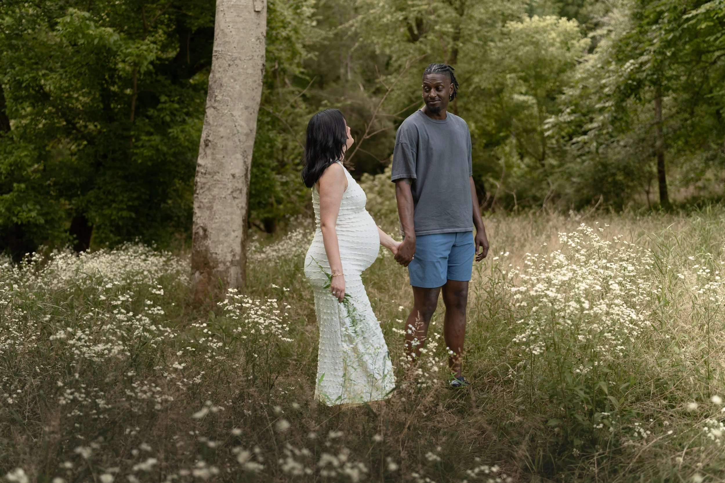 A couple holding hands and walking through a field of small white flowers in a wooded area. The woman is pregnant and wearing a white dress, and the man is wearing a gray T-shirt and blue shorts.