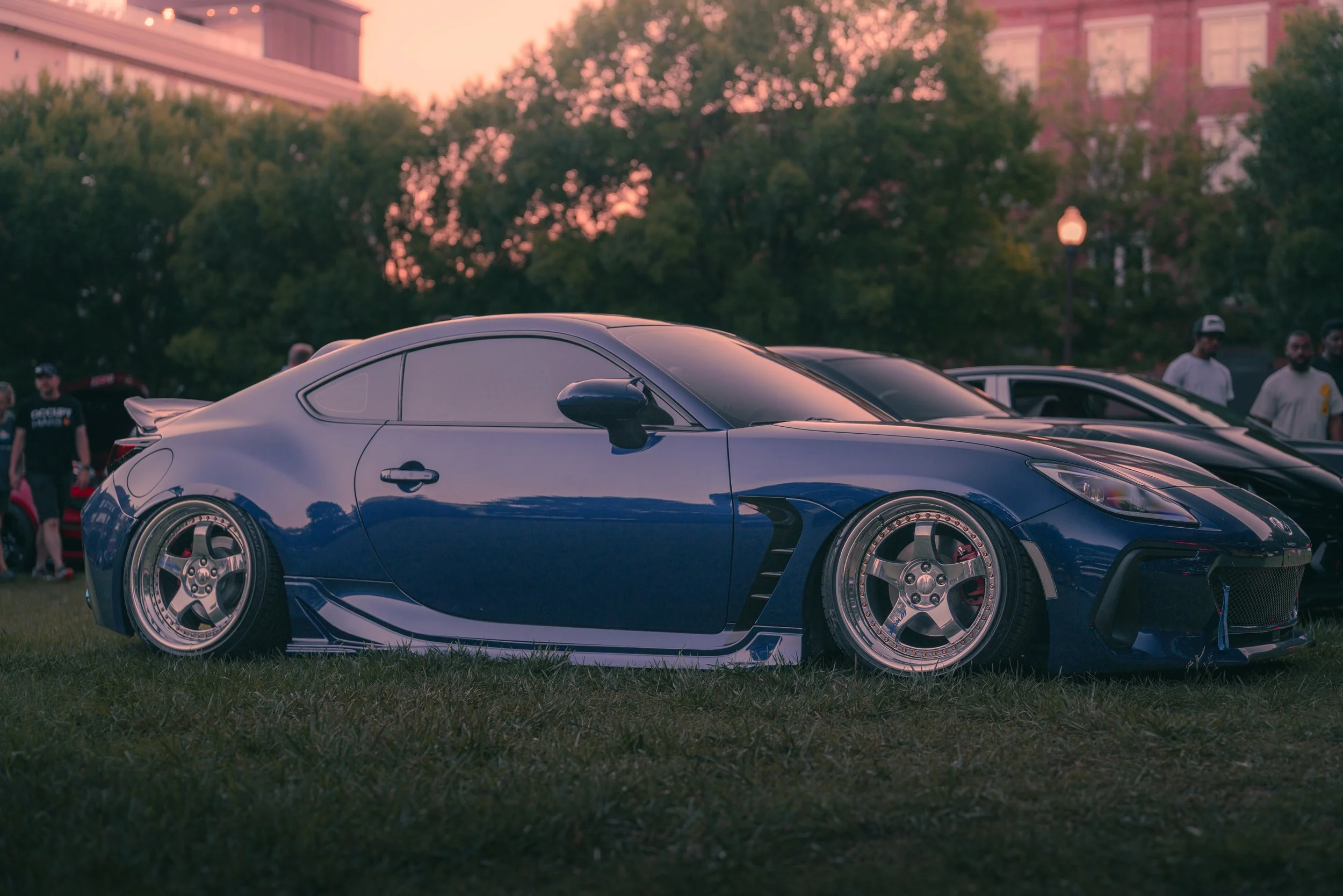 A shiny blue sports car parked on the grass at a car show during sunset with people in the background along with trees and buildings.