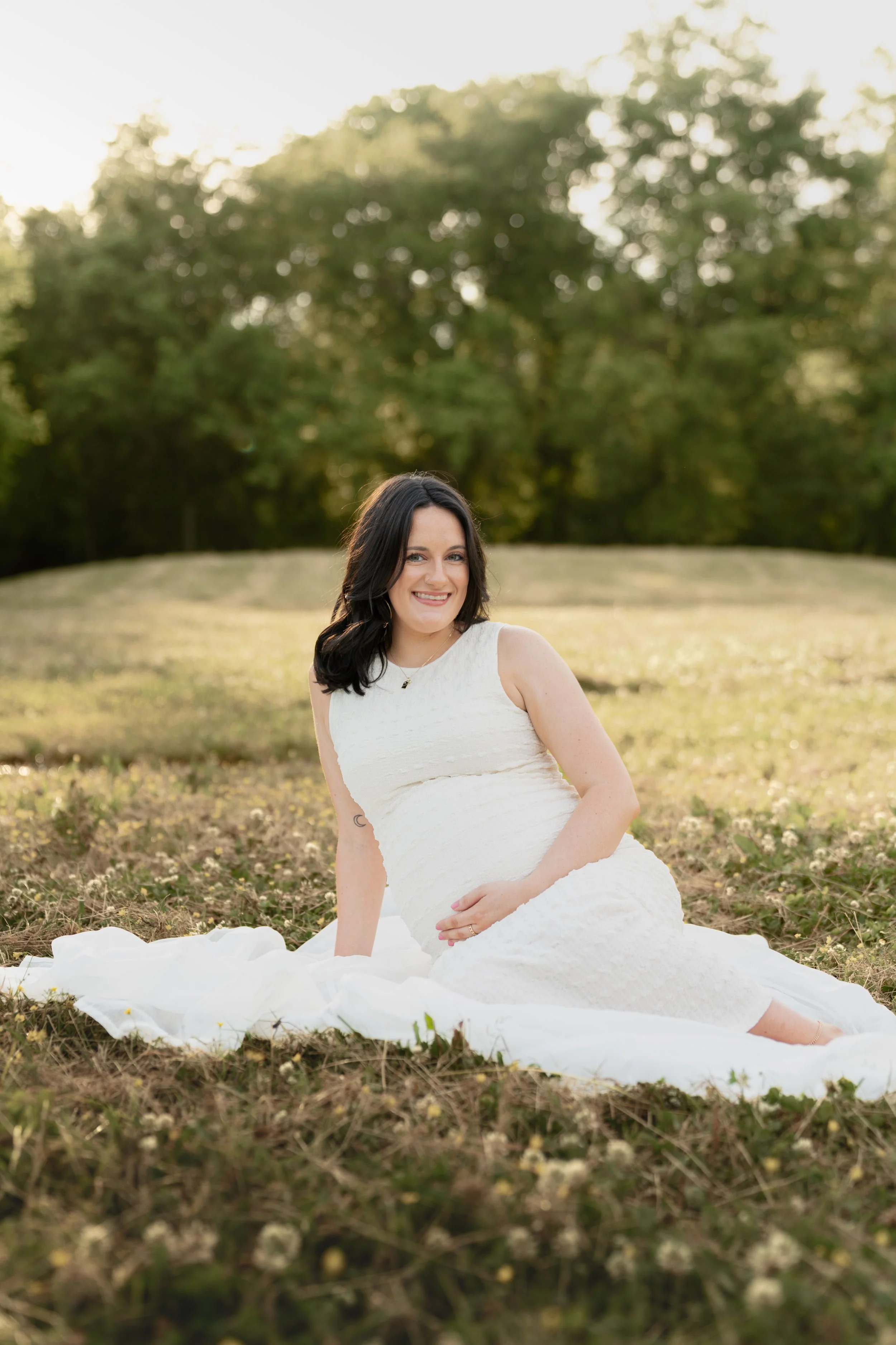 A woman in a white dress with dark hair and a tattoo on her left arm, sitting on grass outdoors with trees in the background, smiling at the camera.