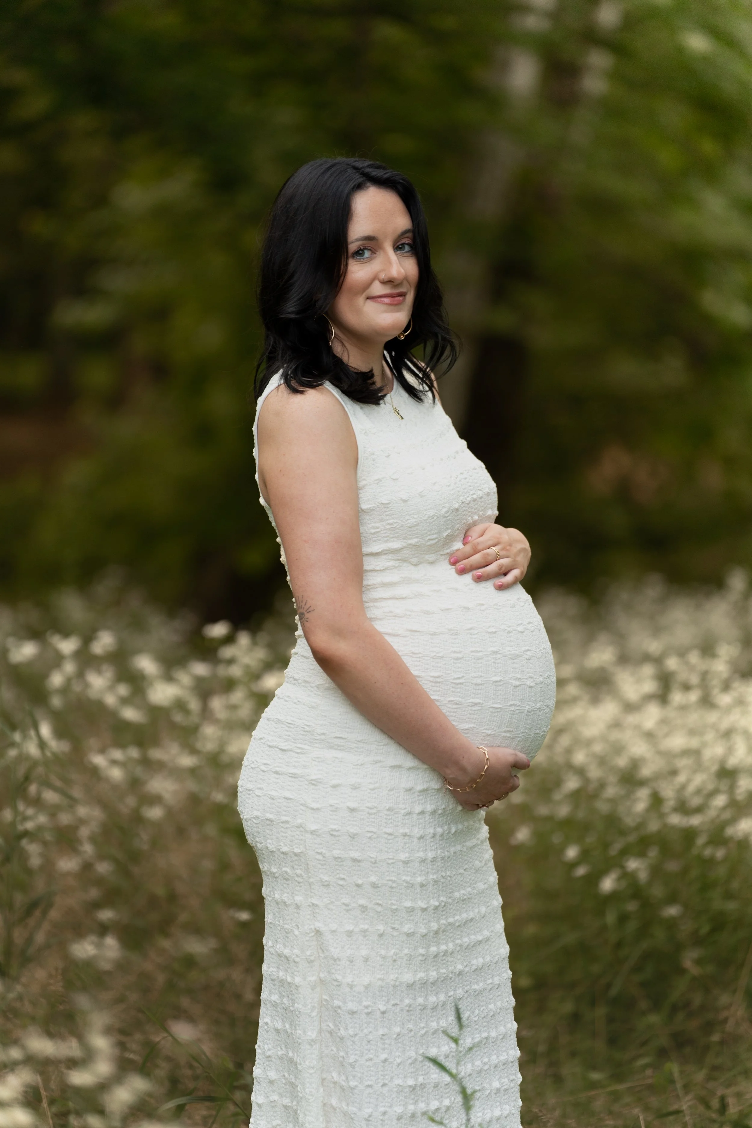 A pregnant woman with black hair and blue eyes wearing a sleeveless white lace dress, standing outdoors in a wooded area during daytime, holding her belly with both hands and smiling softly.