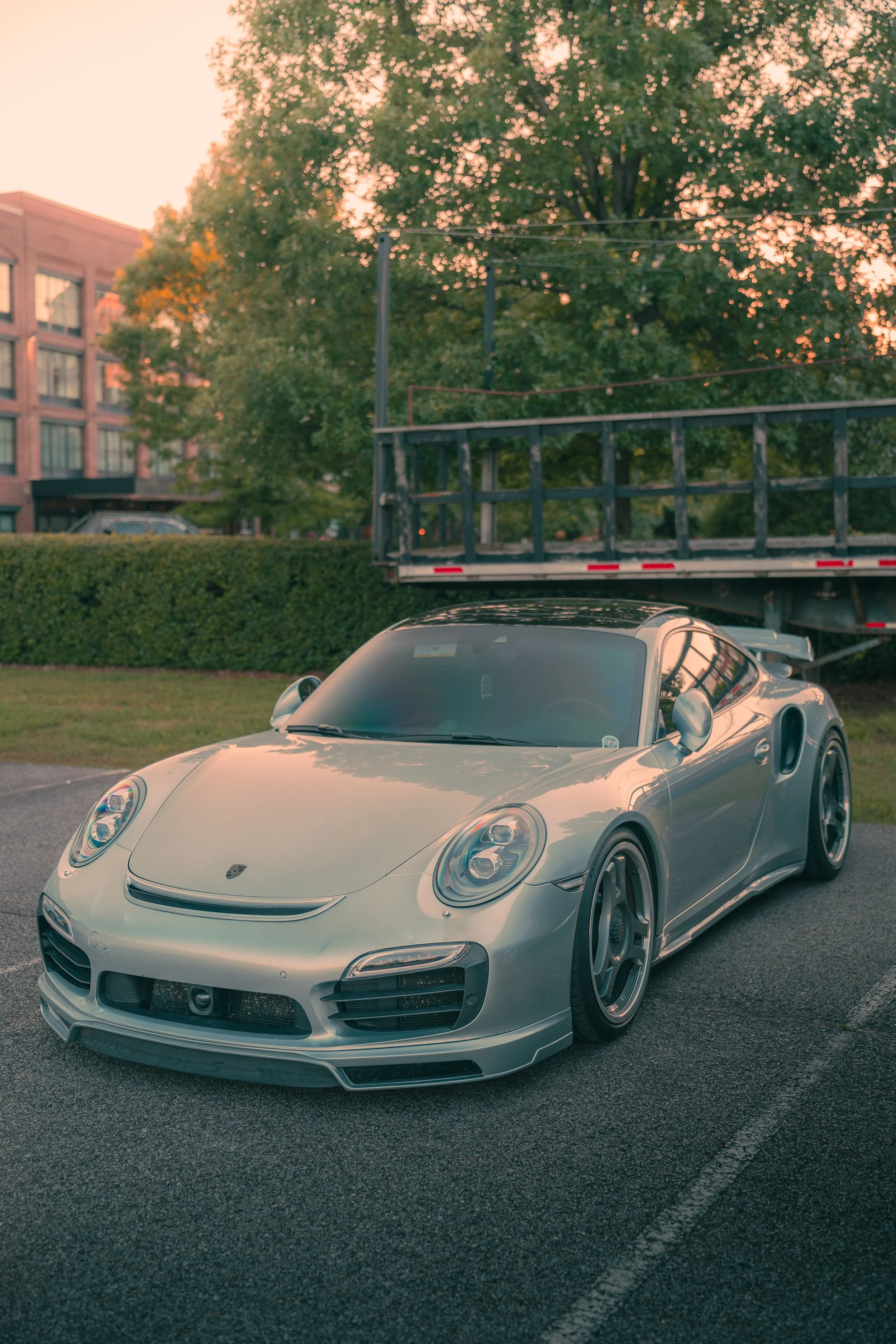A silver Porsche sports car parked in a lot, with a flatbed truck and trees in the background during sunset.