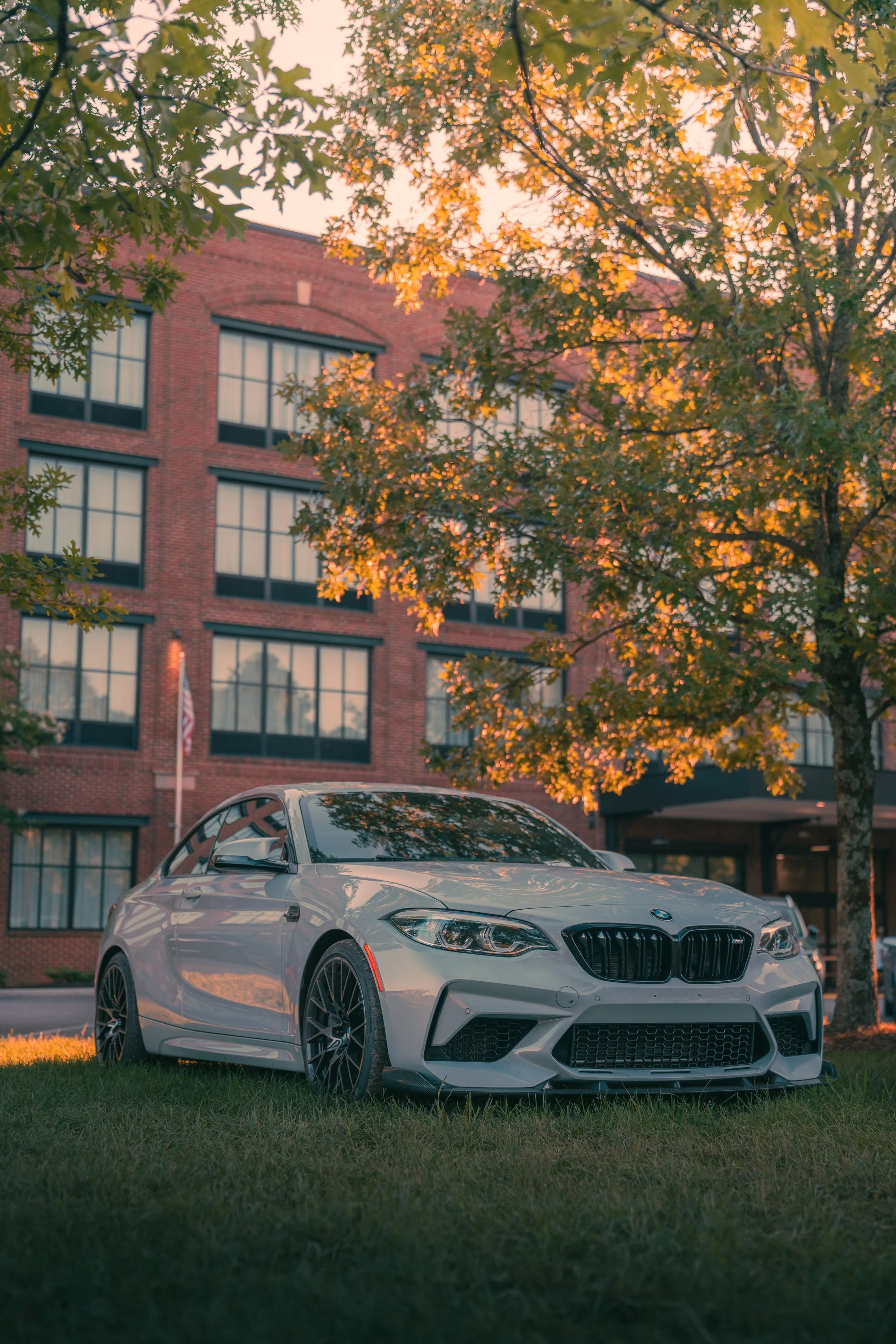 A silver BMW sports car parked on a grassy area in front of a brick apartment building, with trees and sunset in the background.