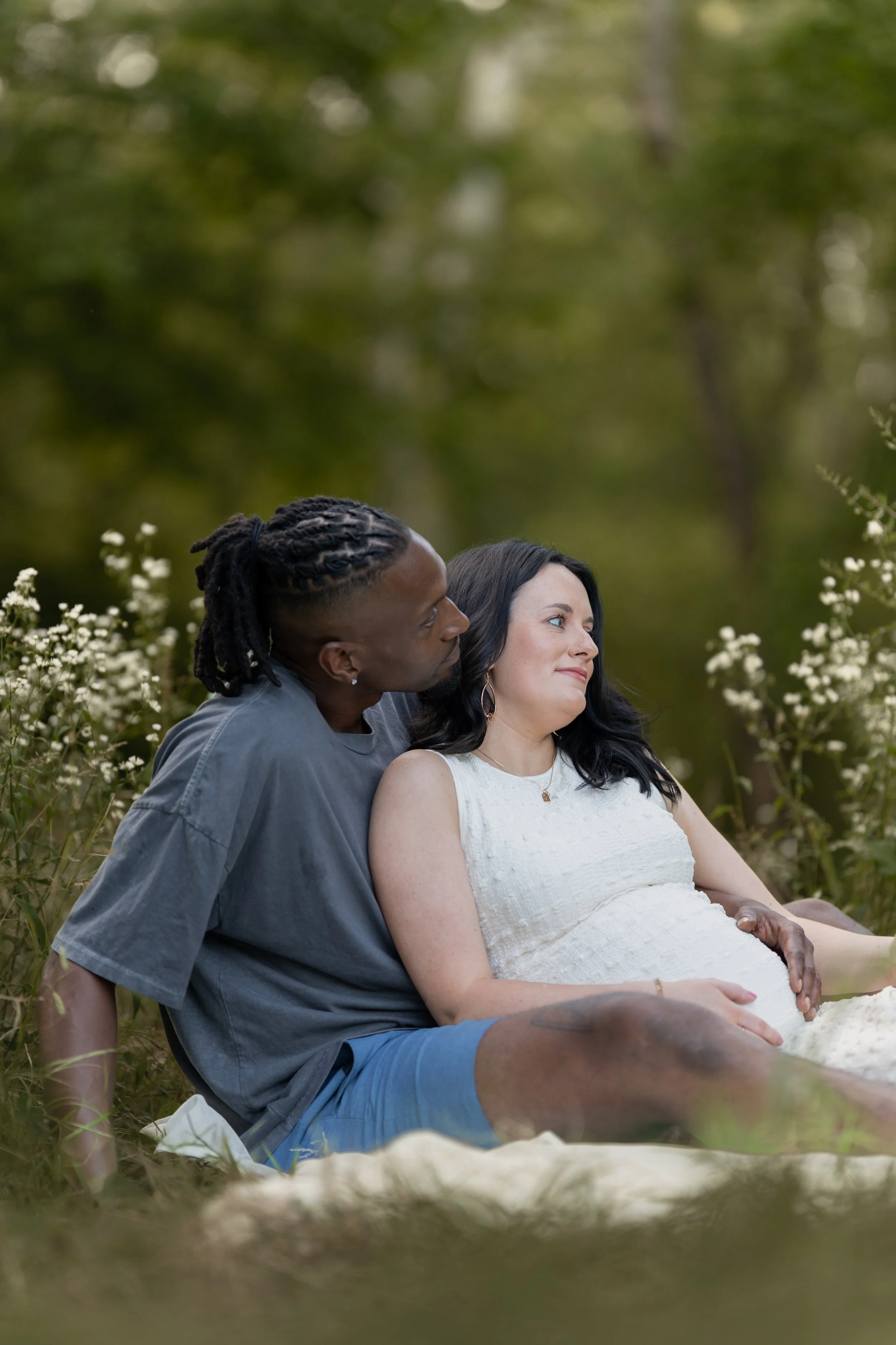A man and a woman sitting outdoors by flowers, with the woman pregnant, sharing a tender moment.