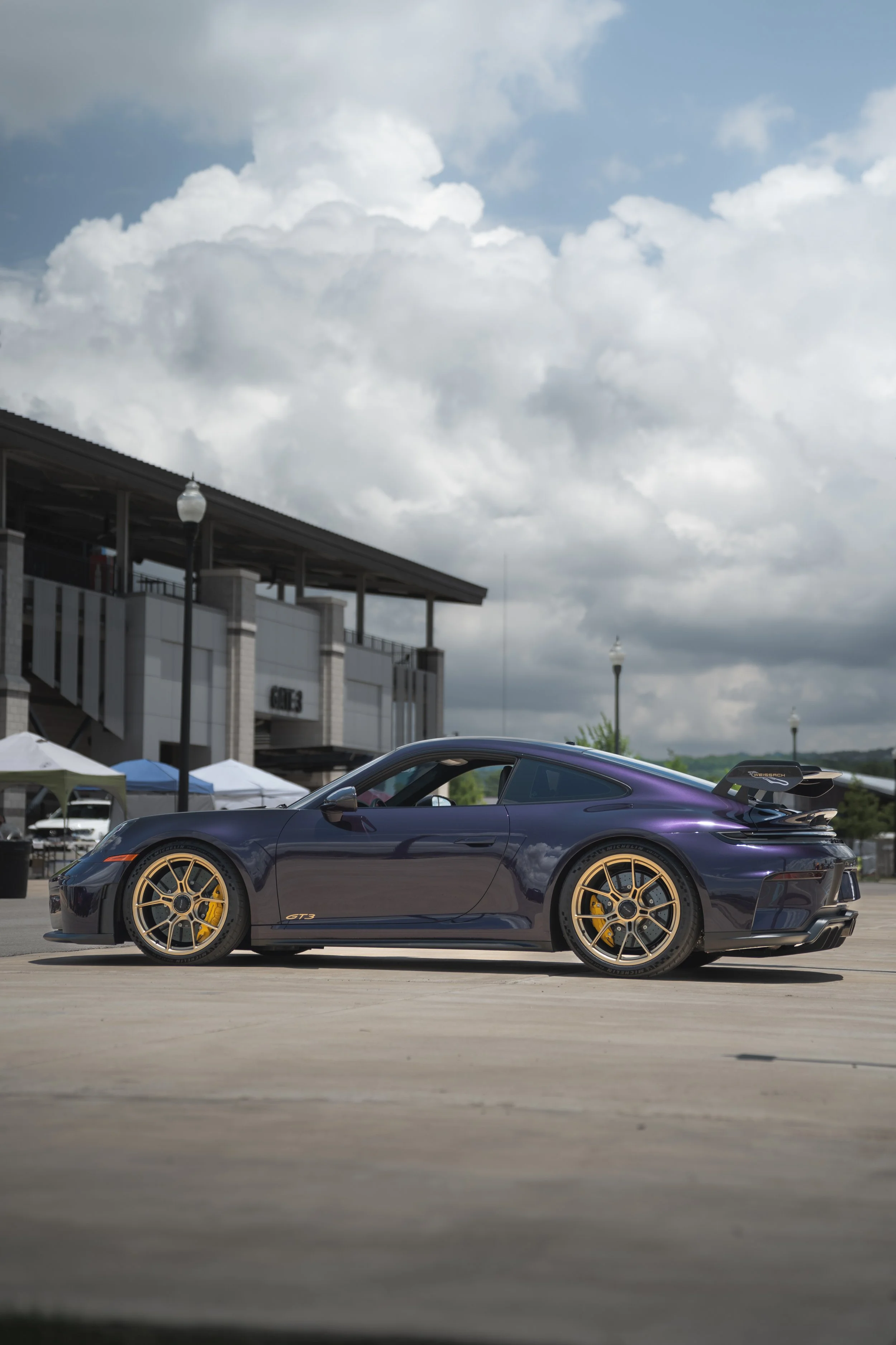 A dark purple sports car with yellow wheels parked on a paved area in front of a building, under a partly cloudy sky.