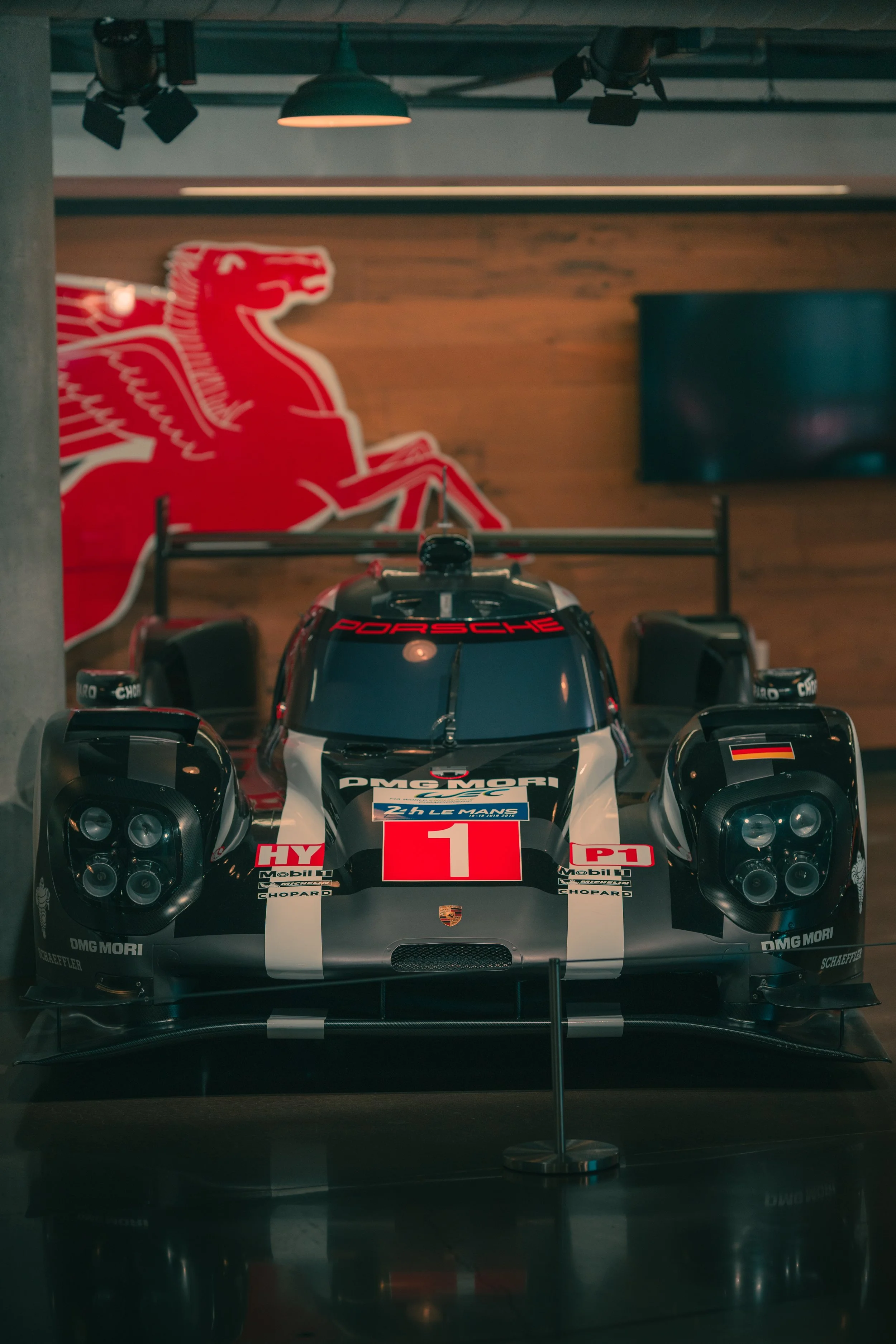A race car displayed behind a glass barrier in a museum, with a large red and white animal logo on the wall and various sponsor logos on the car.