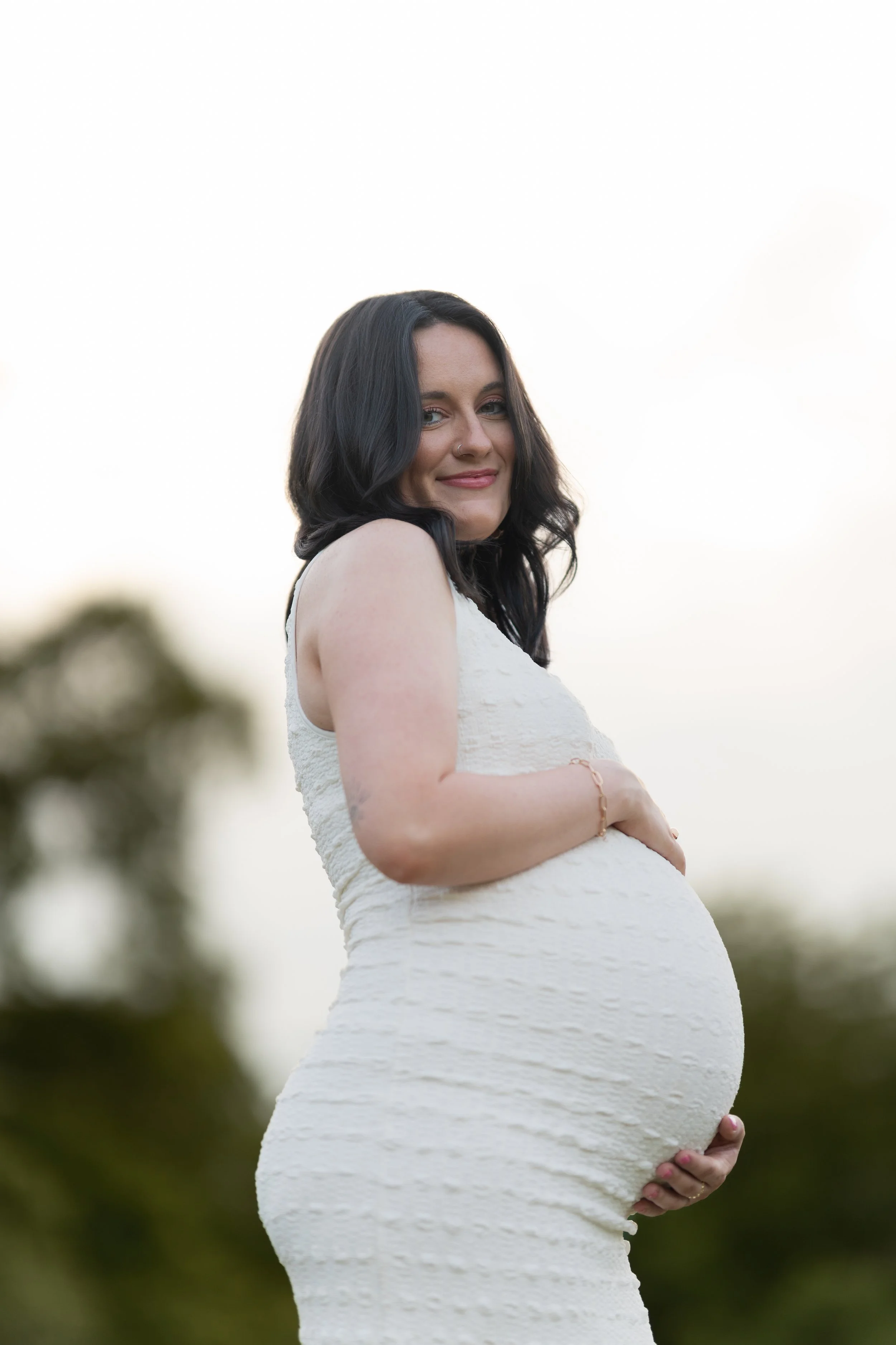 A pregnant woman with dark wavy hair, wearing a white textured sleeveless dress, standing outdoors with a blurred natural background, holding her belly with both hands, smiling softly.
