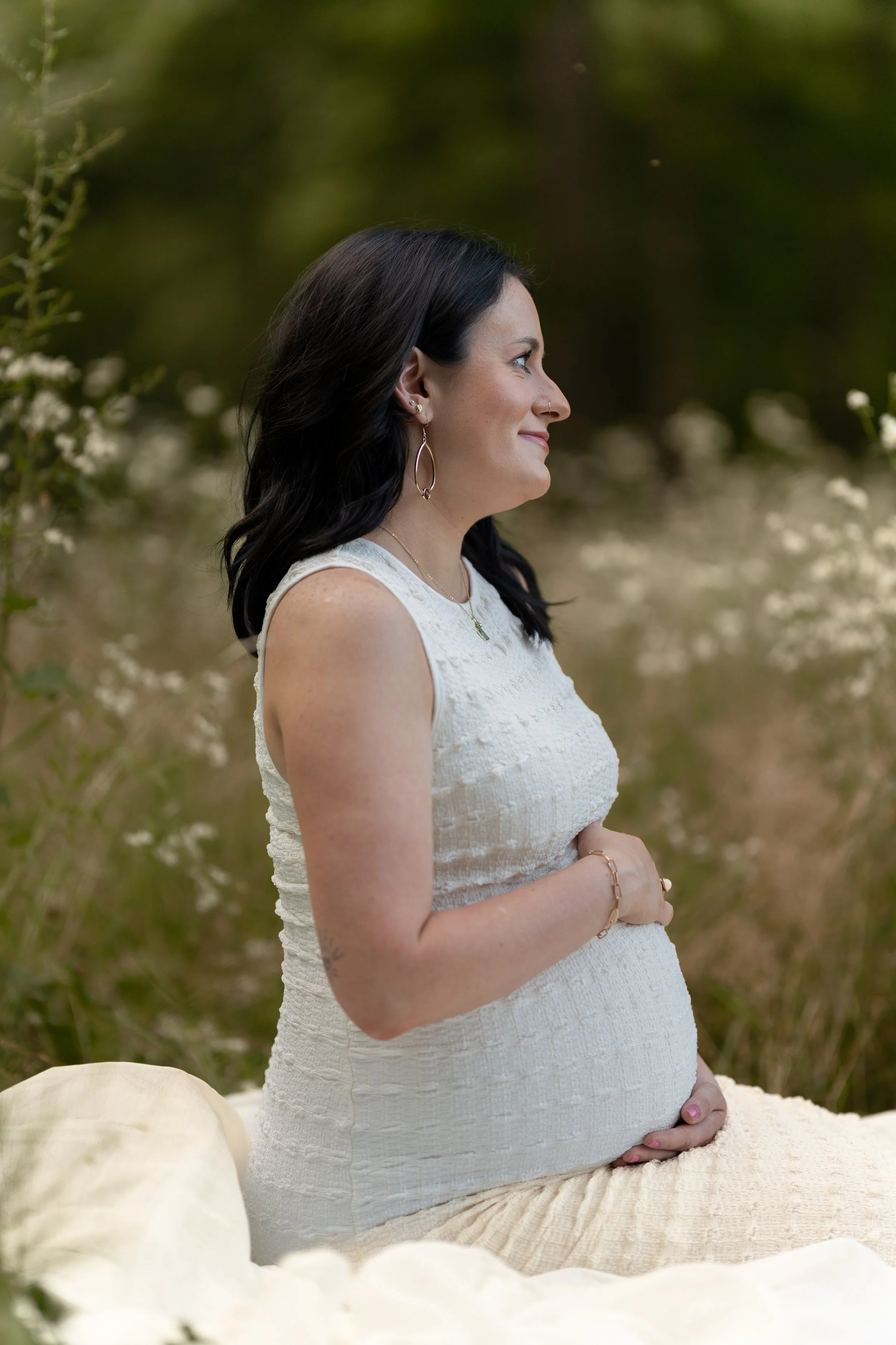 Pregnant woman sitting outdoors on a blanket, with her hands on her belly, wearing a white sleeveless dress, in a natural setting with greenery and small white flowers.