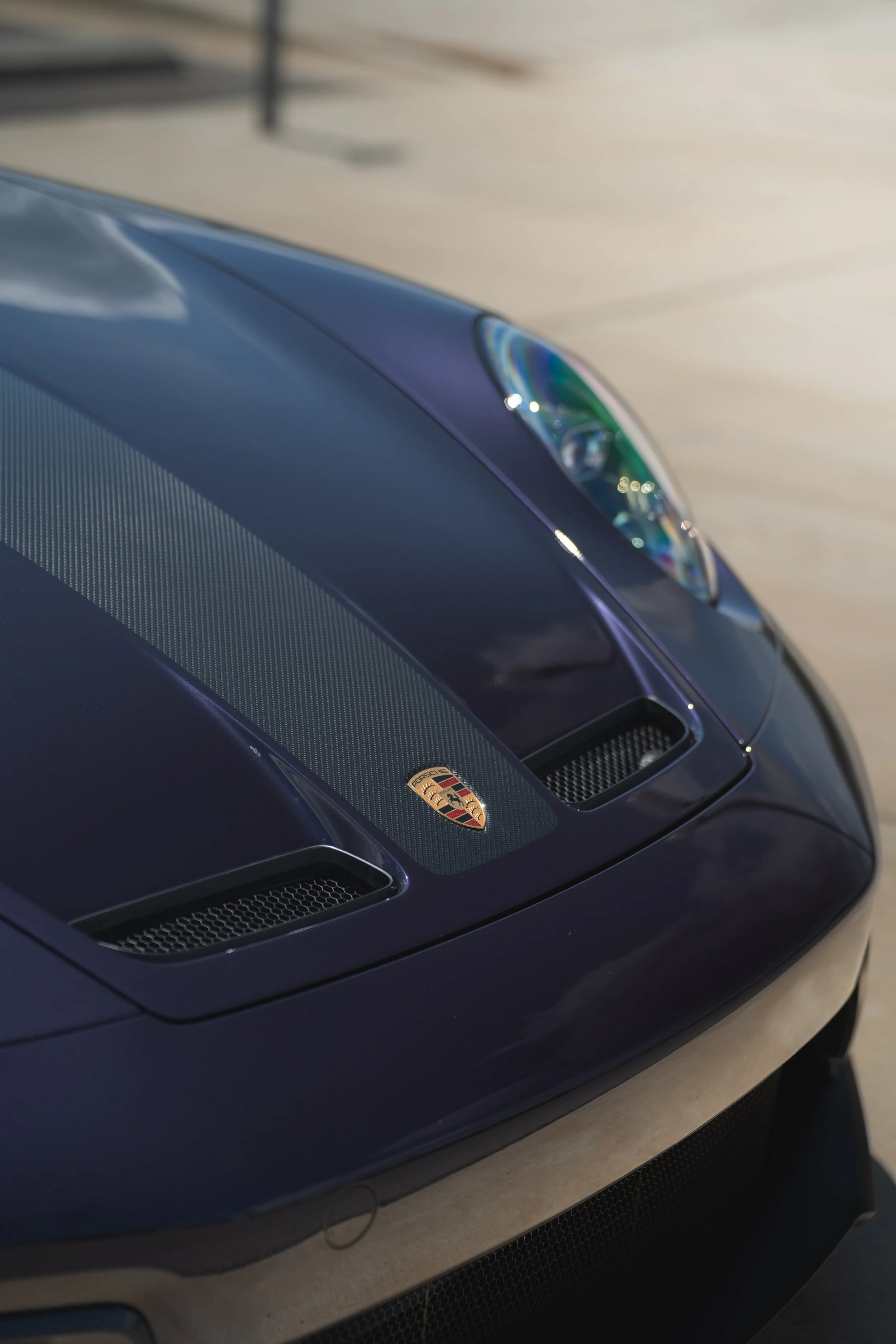 Close-up of the front of a blue Porsche sports car showing the Porsche crest on the hood and the headlight.