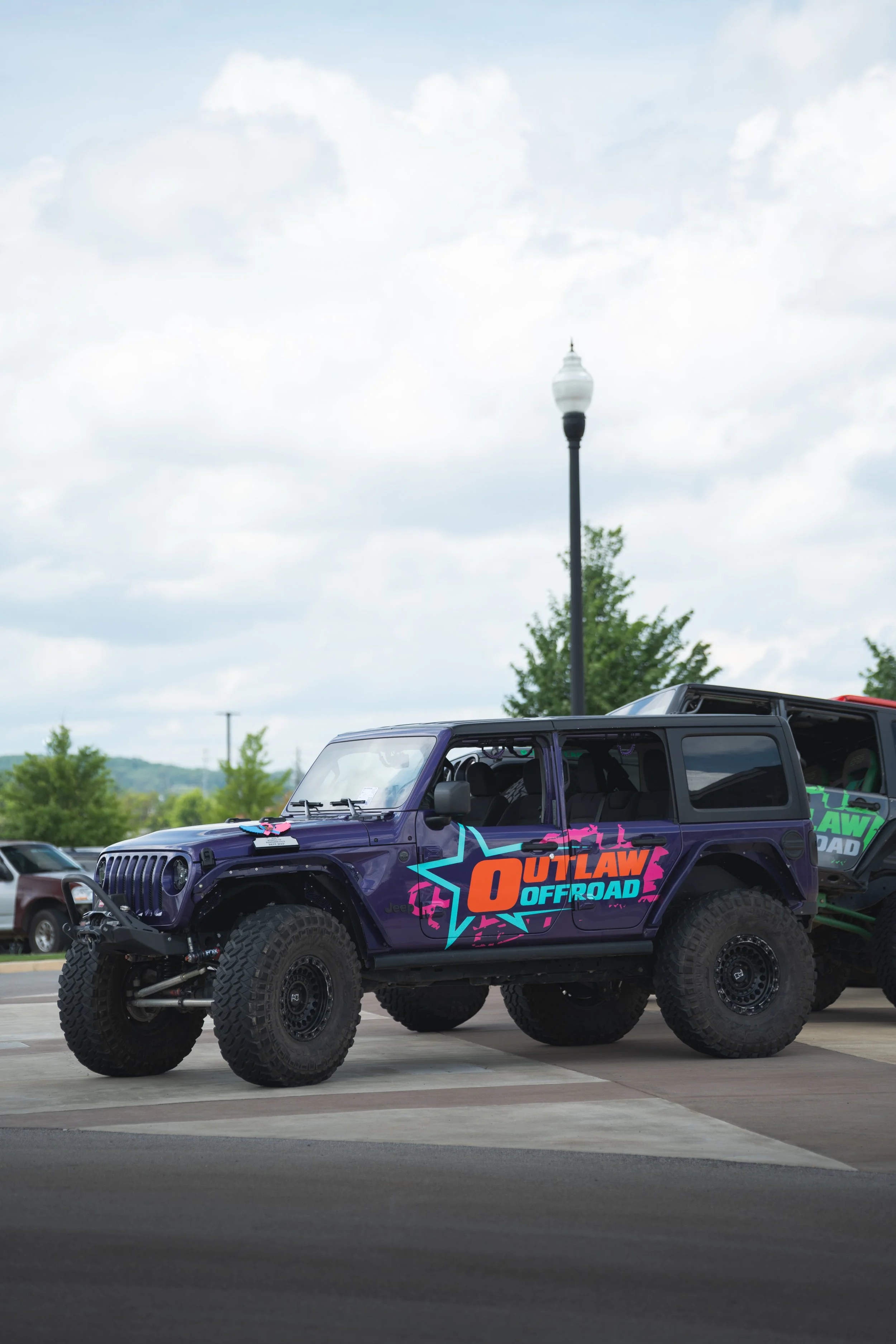 A purple Jeep off-road vehicle with large tires and a colorful decal that reads 'OUTLAW OFFROAD' parked in a lot.