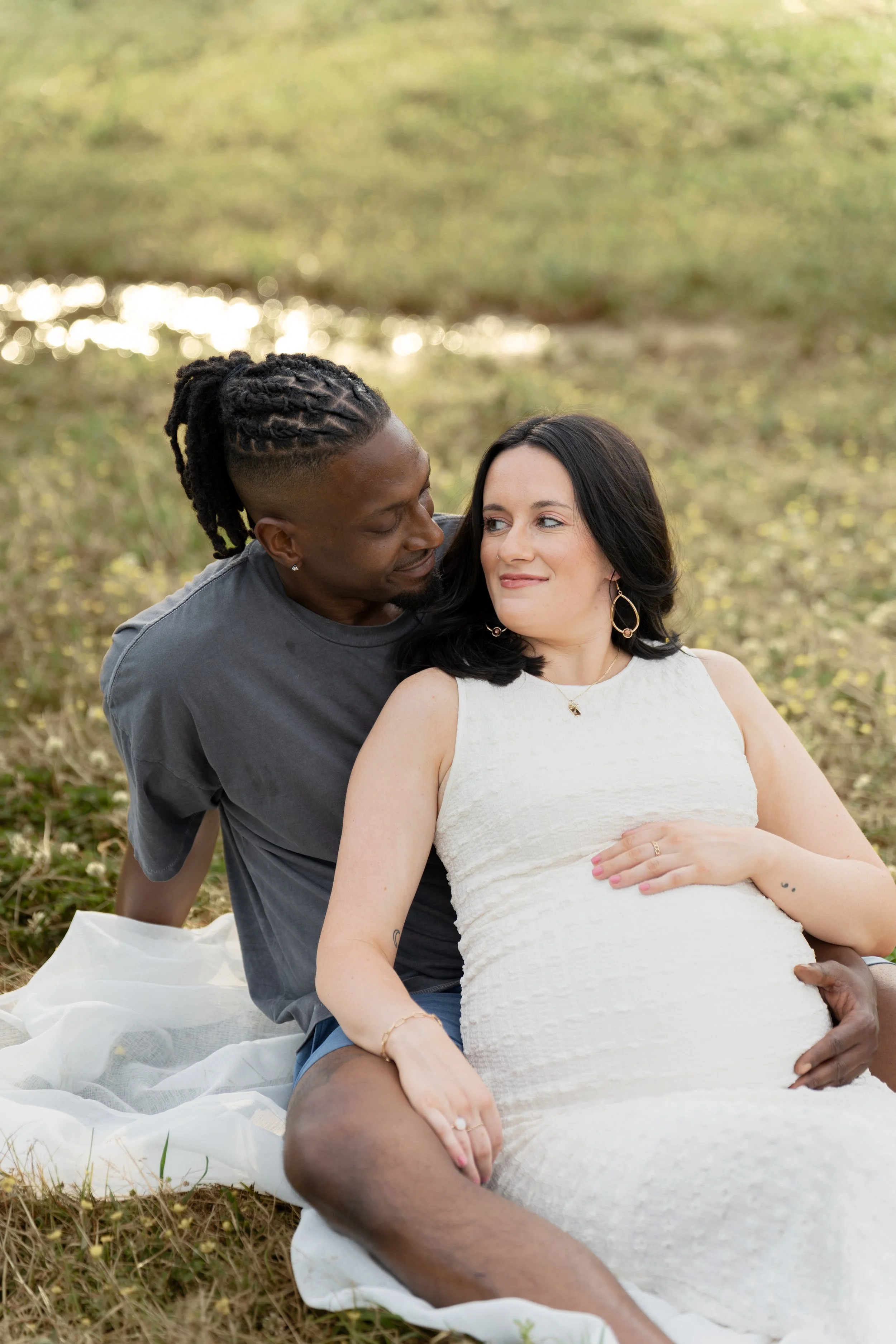 A couple sitting outdoors on a white blanket in a grassy area near a pond. The man has dark skin, dreadlocks, a beard, and is wearing a gray shirt. The woman has light skin, dark hair, and is wearing a white dress and earrings. They are smiling and g