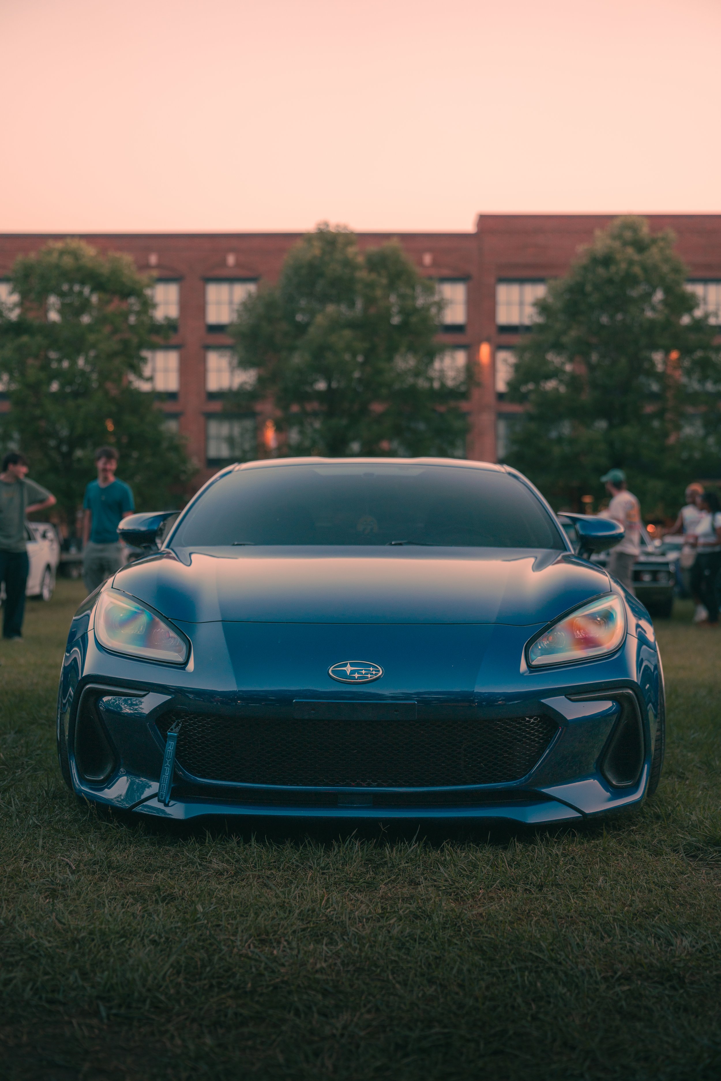 Black sports car with a Subaru emblem in the front, parked on grass at an outdoor event during sunset, with a brick building and trees in the background, and people standing around.