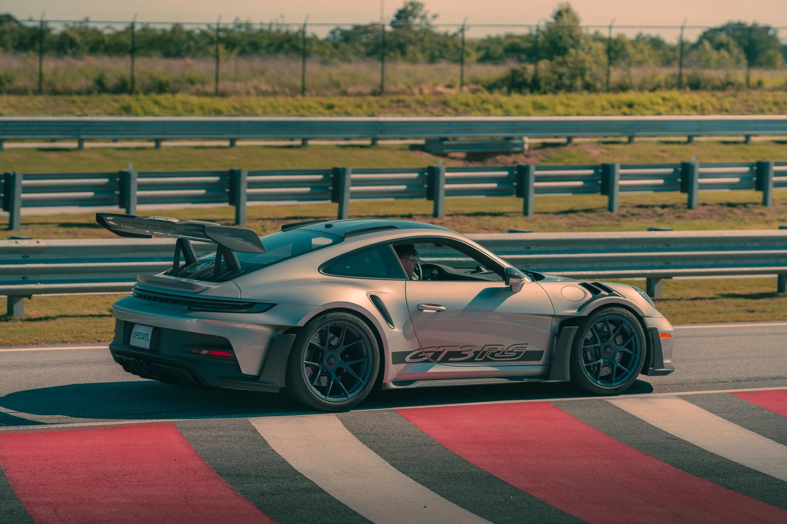 A silver Porsche 911 GT3 RS race car on a race track, with a large rear wing, black wheels, and a driver inside, next to a curbed section marked with red and white paint.