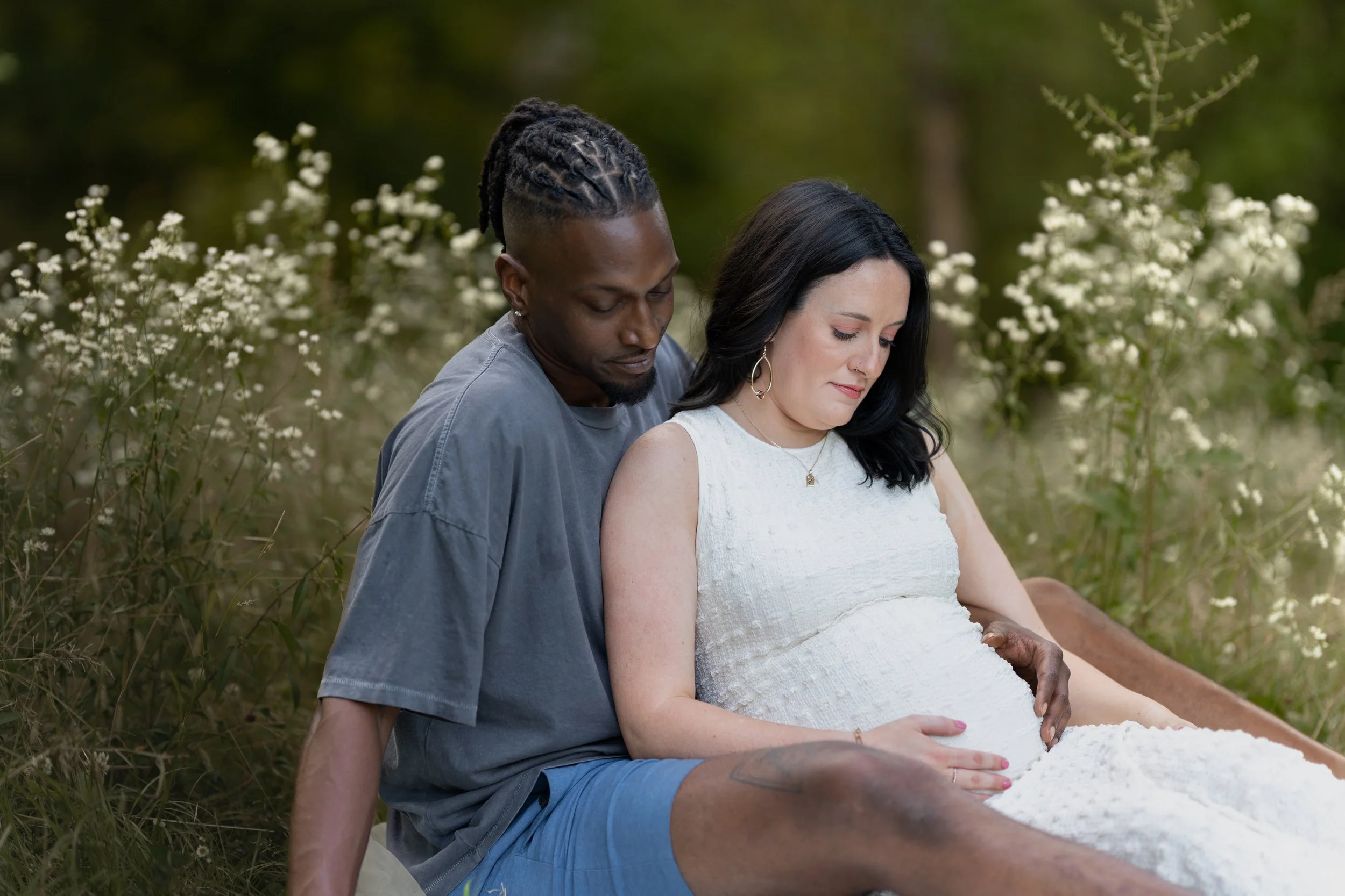 A man and a pregnant woman sitting outdoors in a field of white flowers, with trees in the background, both looking down peacefully with eyes closed.