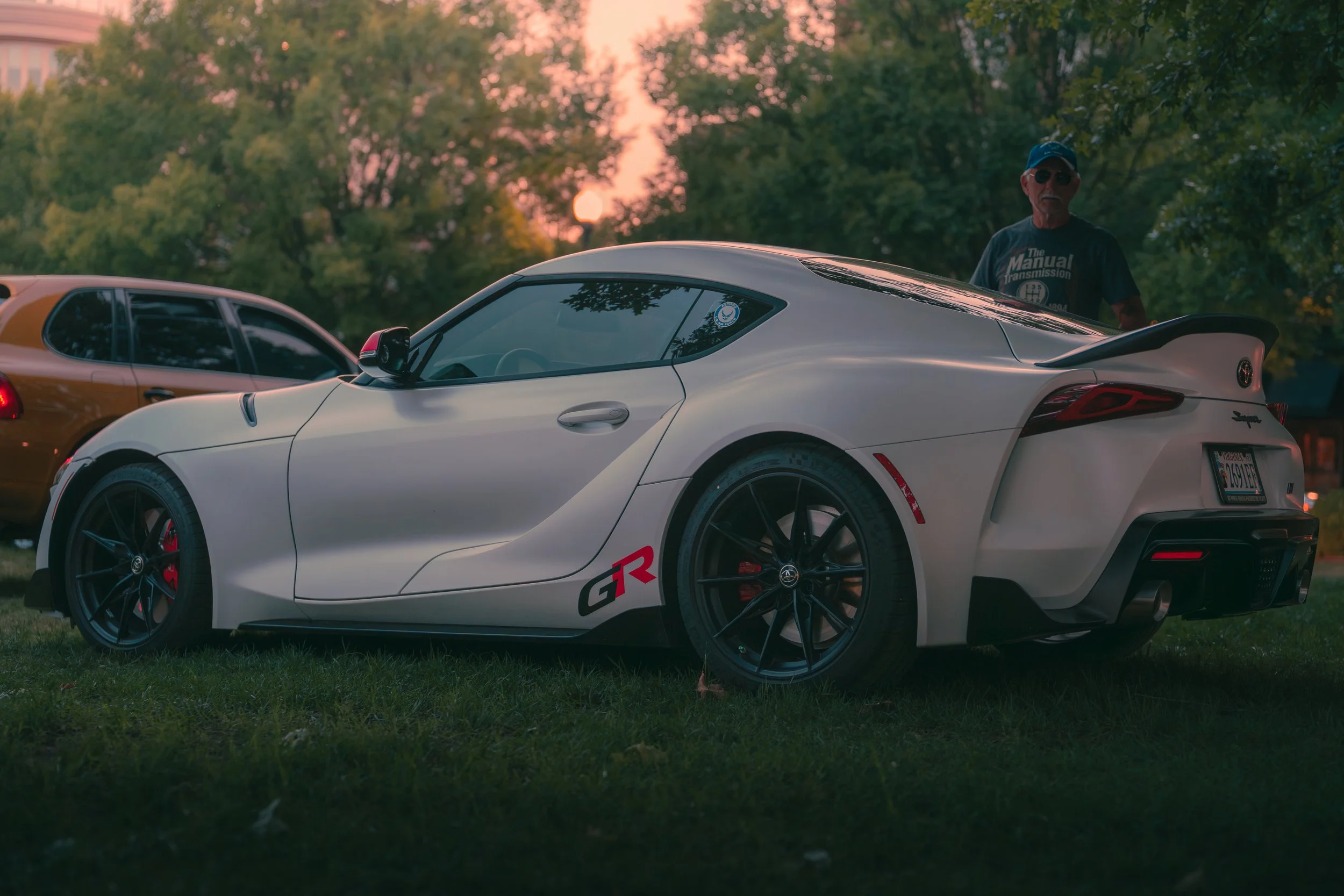 A white sports car with a black spoiler and RedGR decal parked on grass, with a man in sunglasses and a cap standing beside it during sunset in a park with trees in the background.