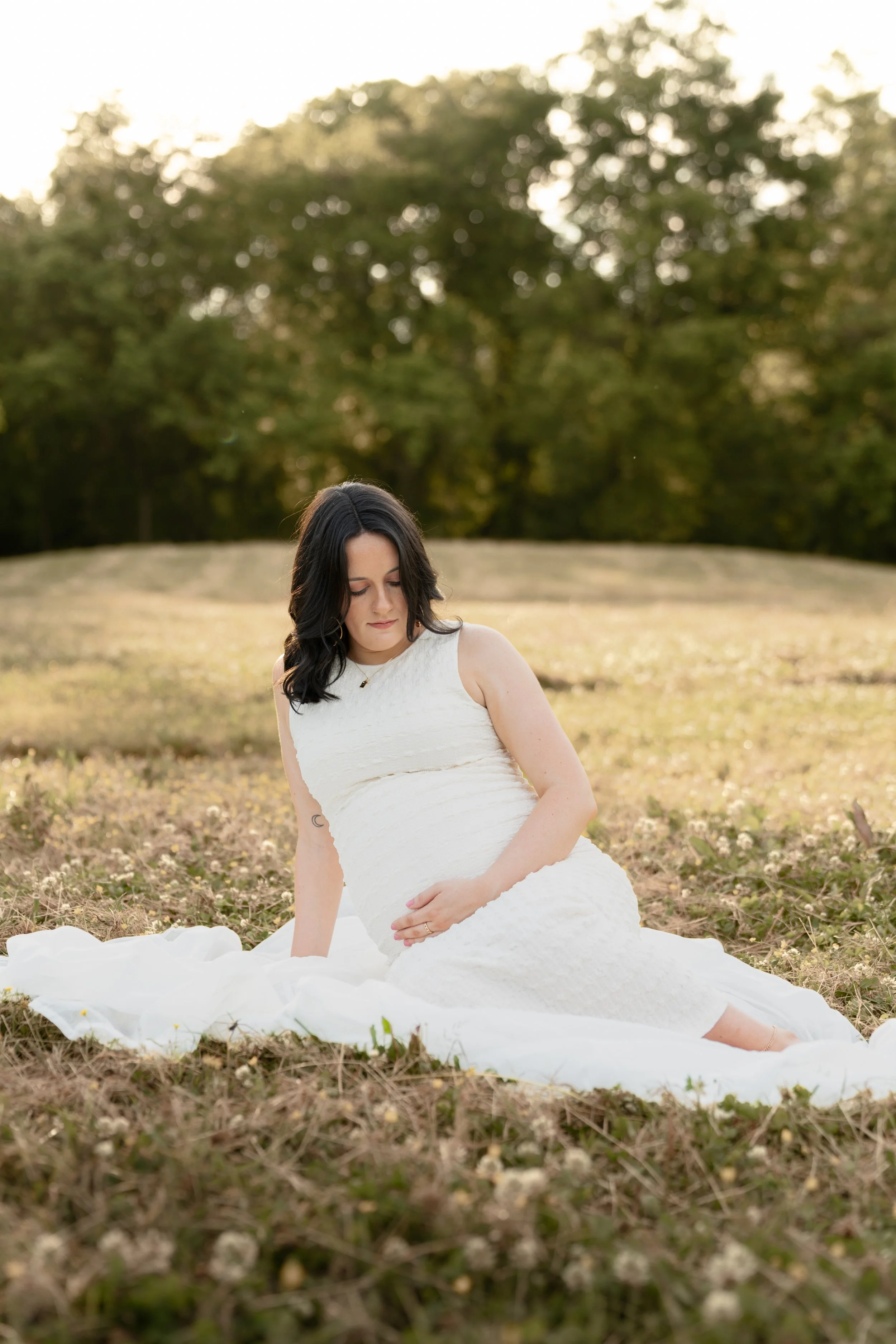 A woman in a white dress sitting on the ground outdoors during sunset or late afternoon, with trees in the background.