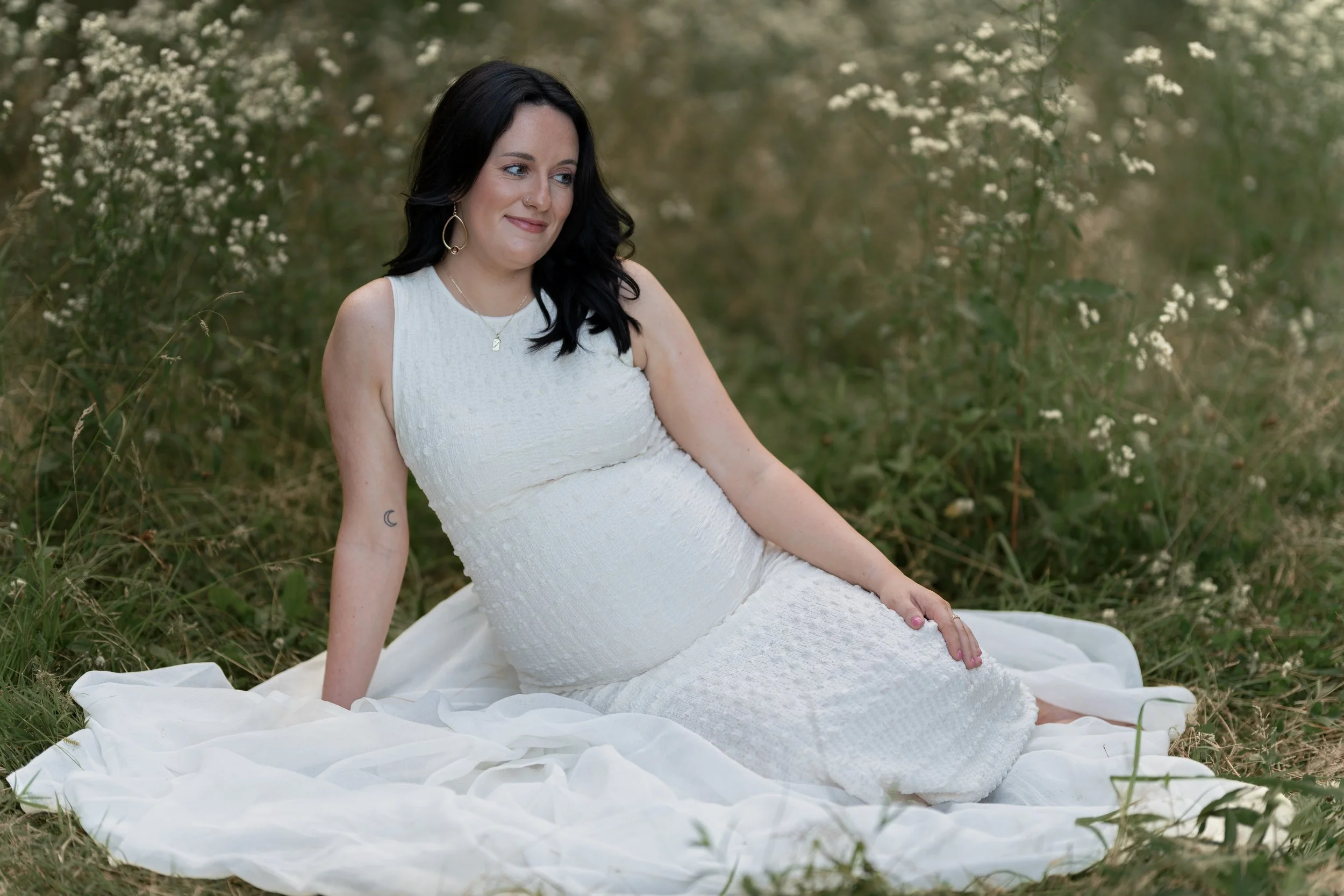 A woman in a white dress sitting on a white cloth in a grassy field surrounded by small white flowers, with dark hair and jewelry, smiling softly.