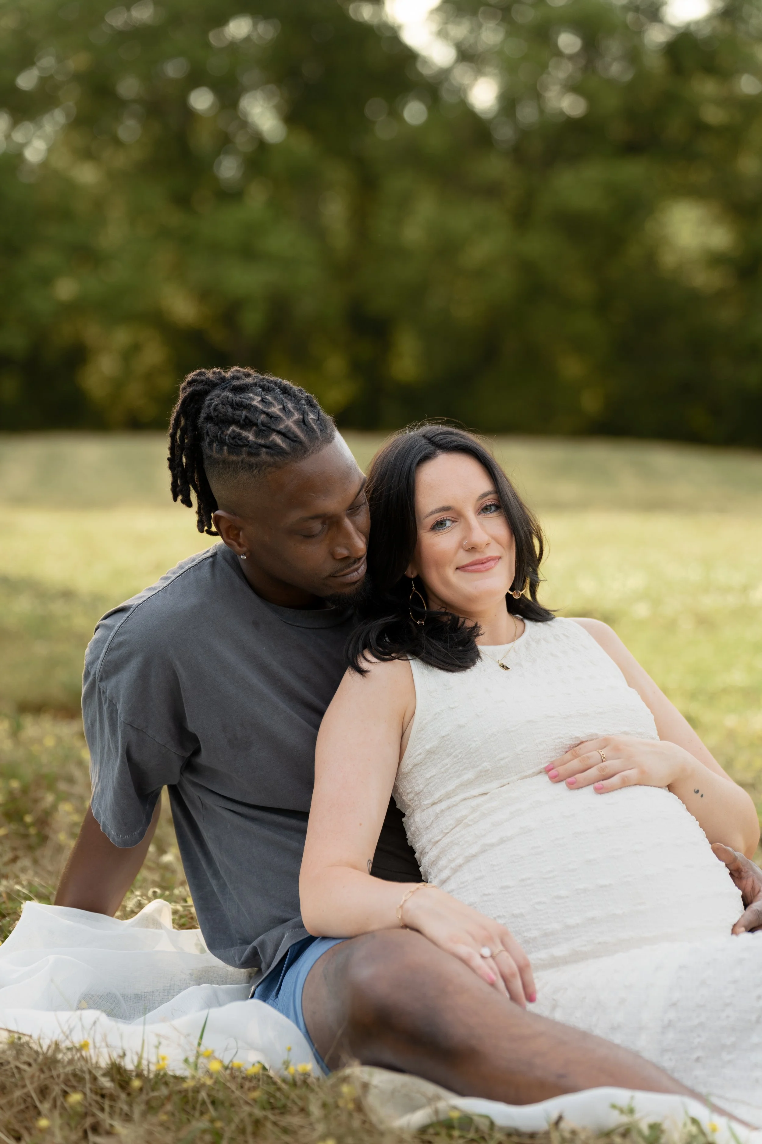 A man and woman sitting on the grass outdoors, with the woman pregnant, in a park or green space during daytime.