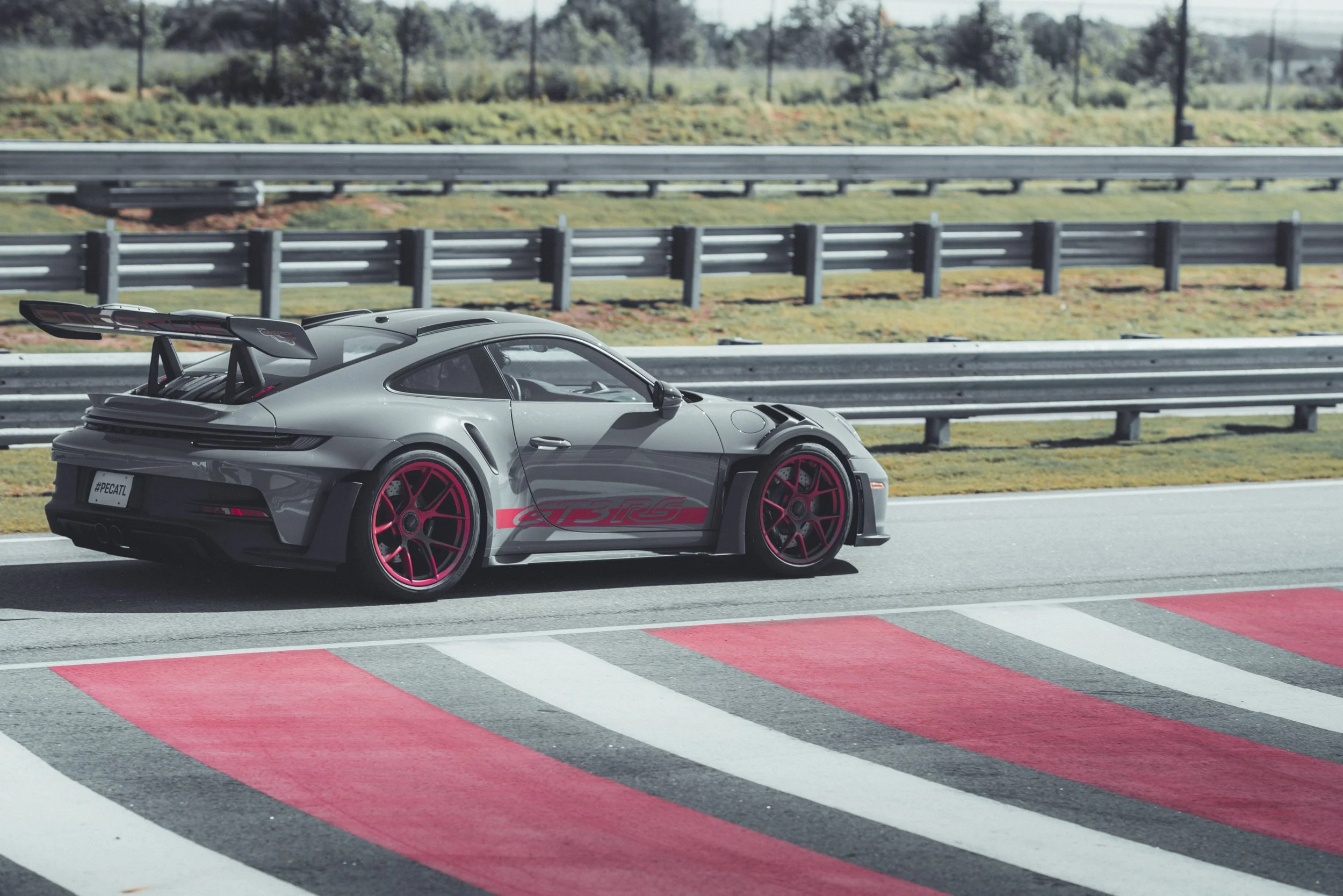 A sleek gray sports car with red wheel accents and a large rear spoiler driving on a race track with painted red, gray, and white sections, alongside a metal guardrail and green landscape in the background.