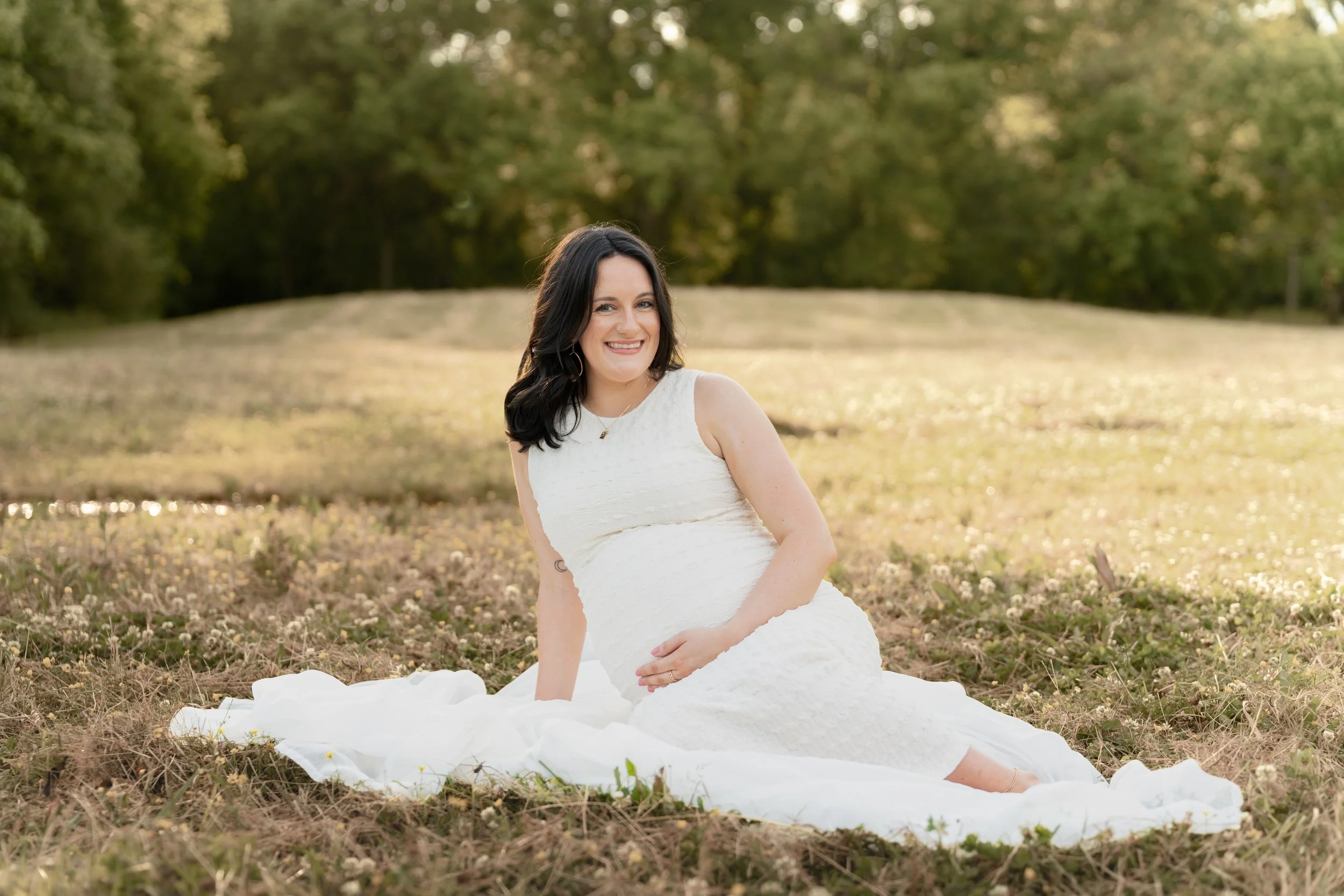 A woman in a white dress sitting on the ground in a field of grass and small flowers, smiling at the camera, with trees in the background.